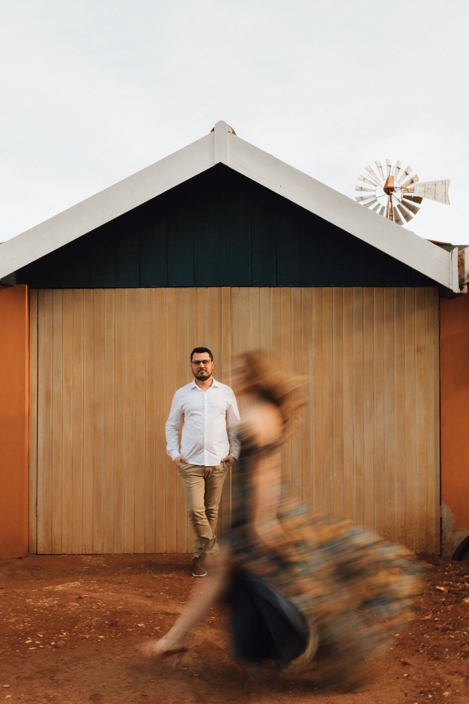 Man in white shirt stands before a wooden barn as a woman in floral dress twirls in motion blur.