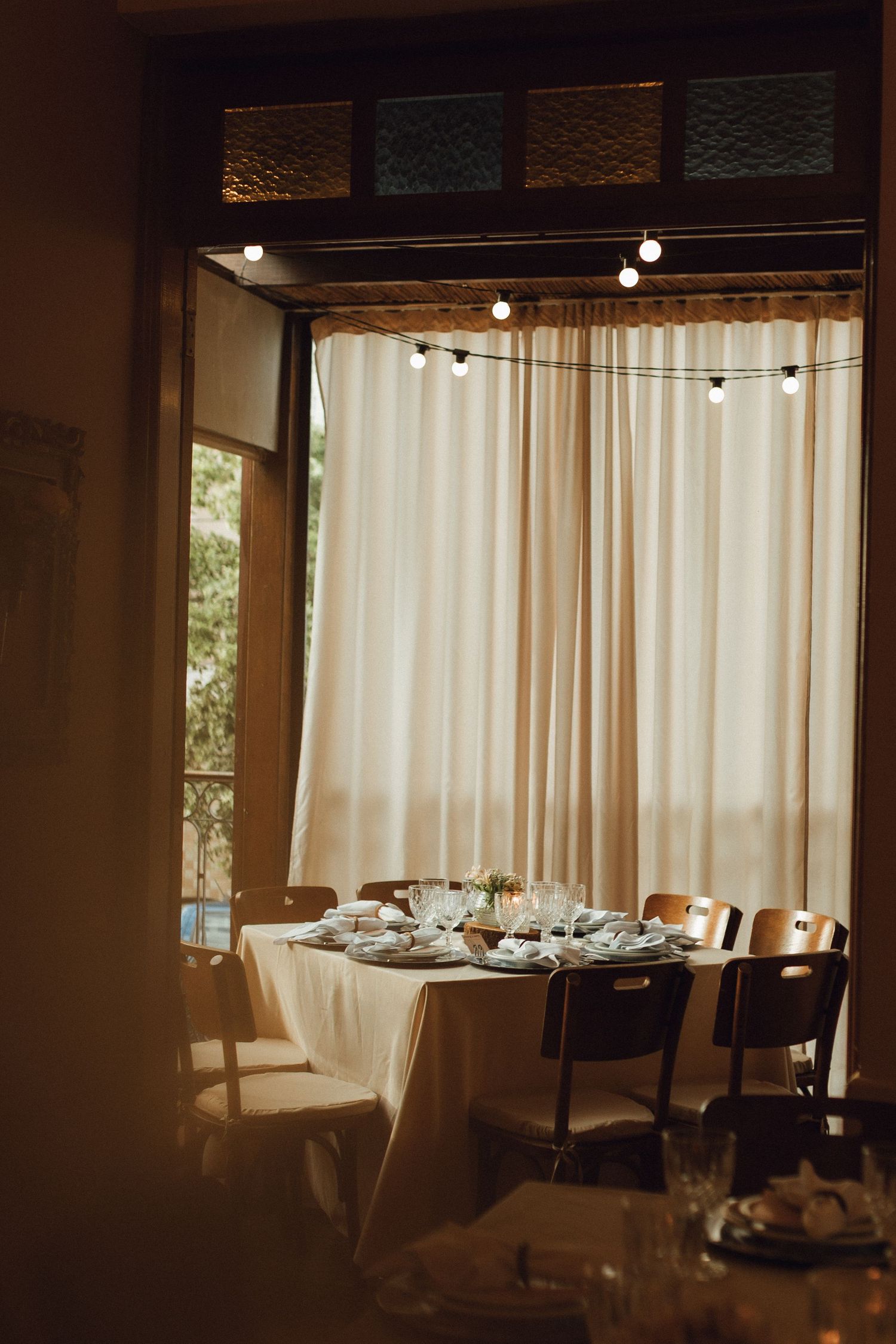 Intimate wedding reception table set with white linens and string lights by sheer curtains.