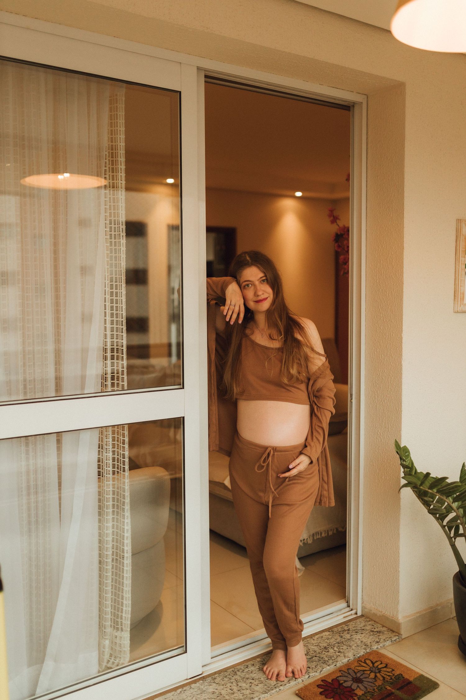 Smiling pregnant woman leans against glass door in brown outfit on apartment balcony.