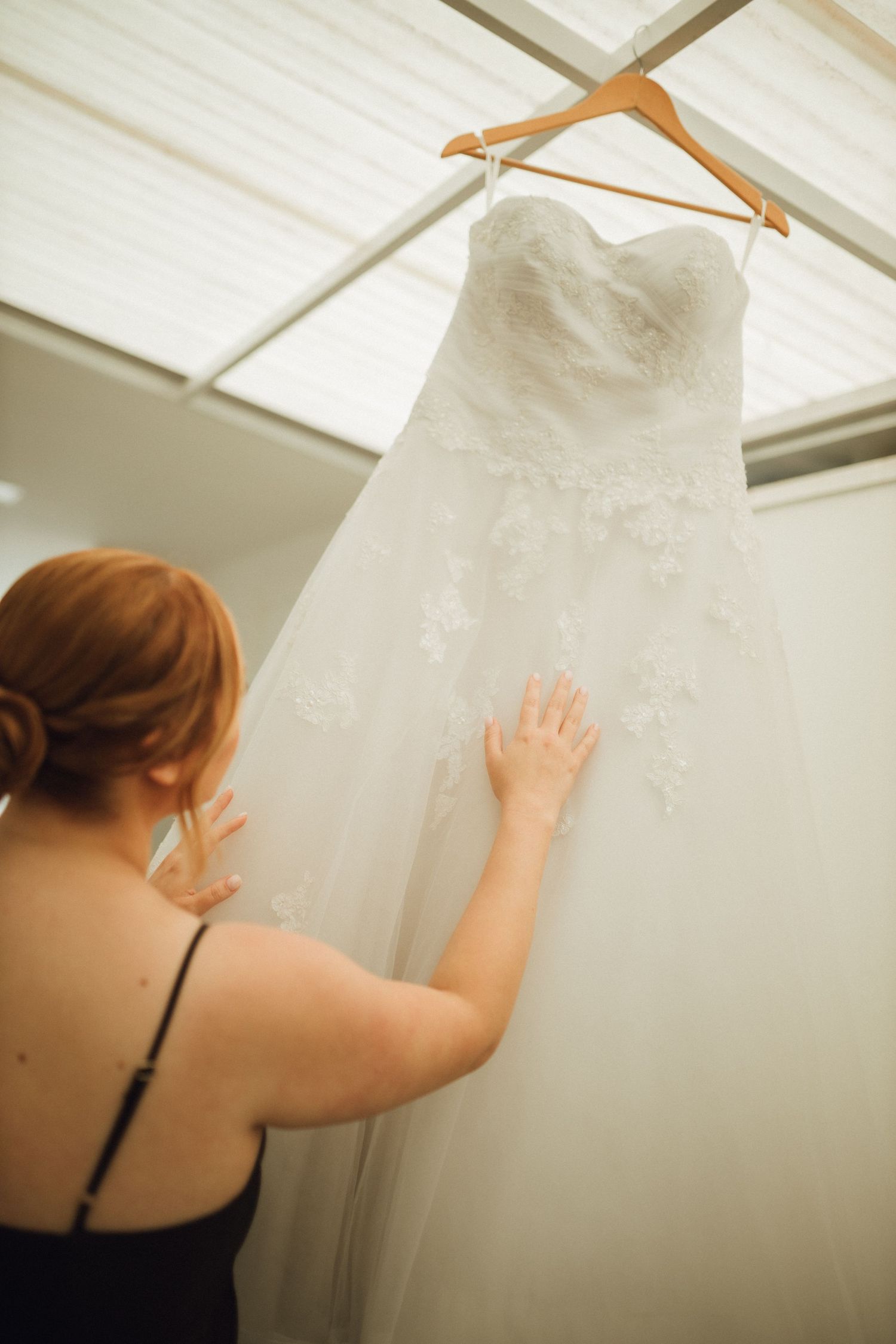 Bride touching elegant white lace strapless wedding gown hanging on wooden hanger in bright room.
