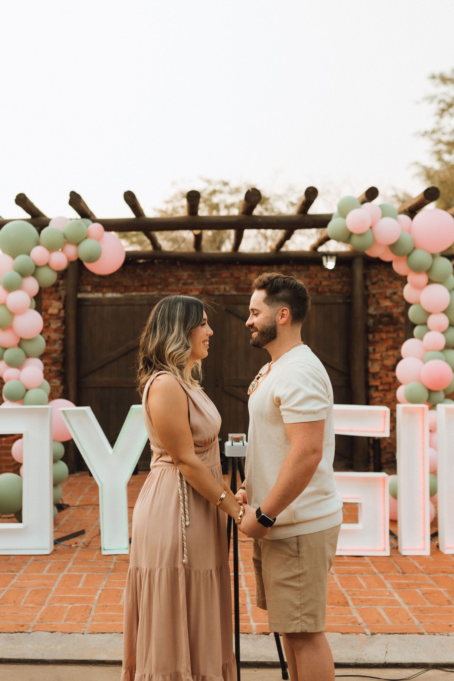 Smiling couple holding hands at gender reveal party with boy or girl balloon letters behind them.
