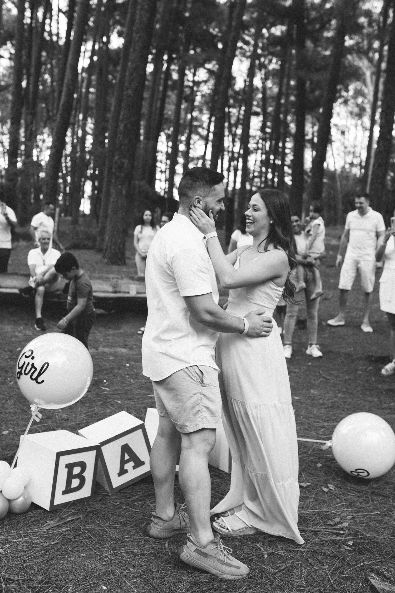 Black and white photo of a couple embracing at a gender reveal party with baby blocks and girl balloon in forest.