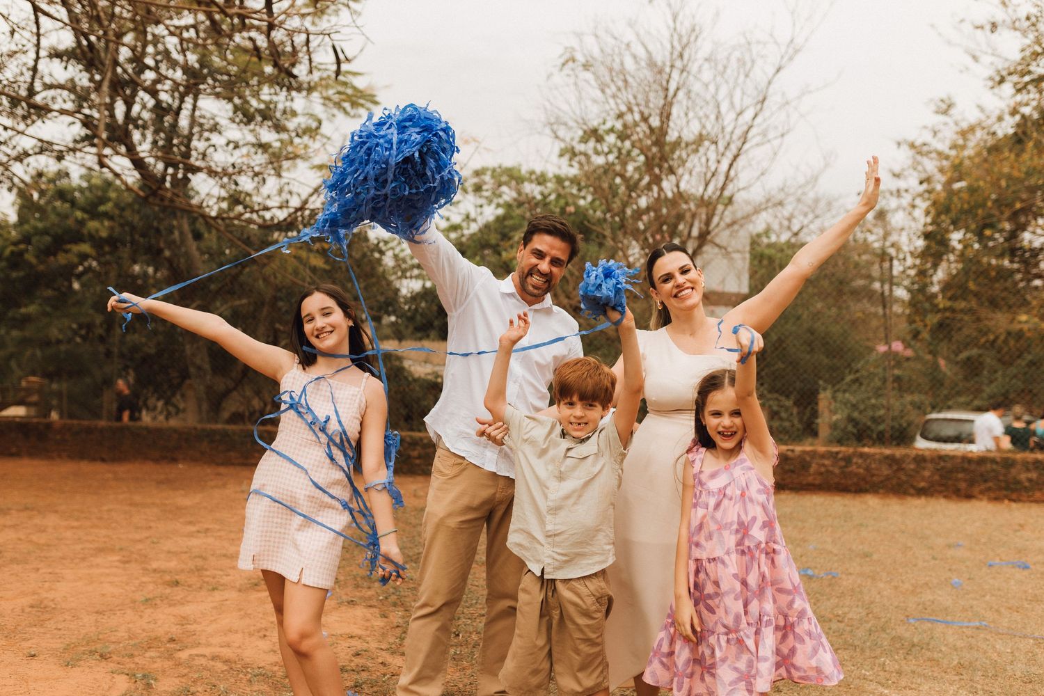 Happy family of five celebrating outdoors with blue pompoms, smiling and raising arms in a sunny park setting.