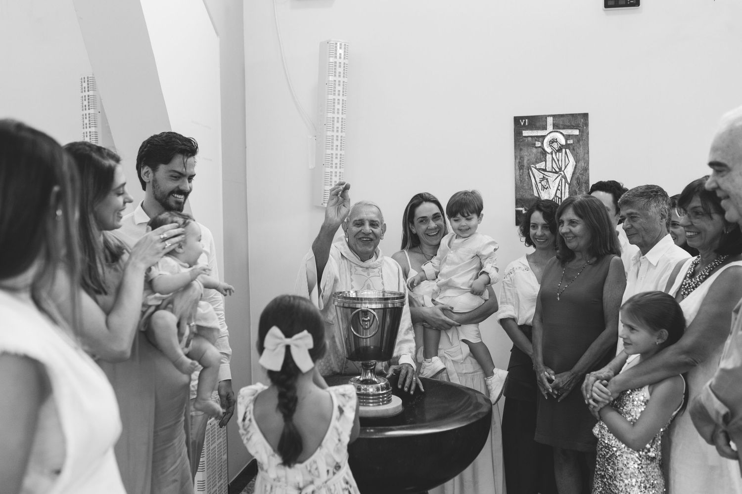 Family gathered around baptismal font during christening ceremony in white church, priest raising hand.