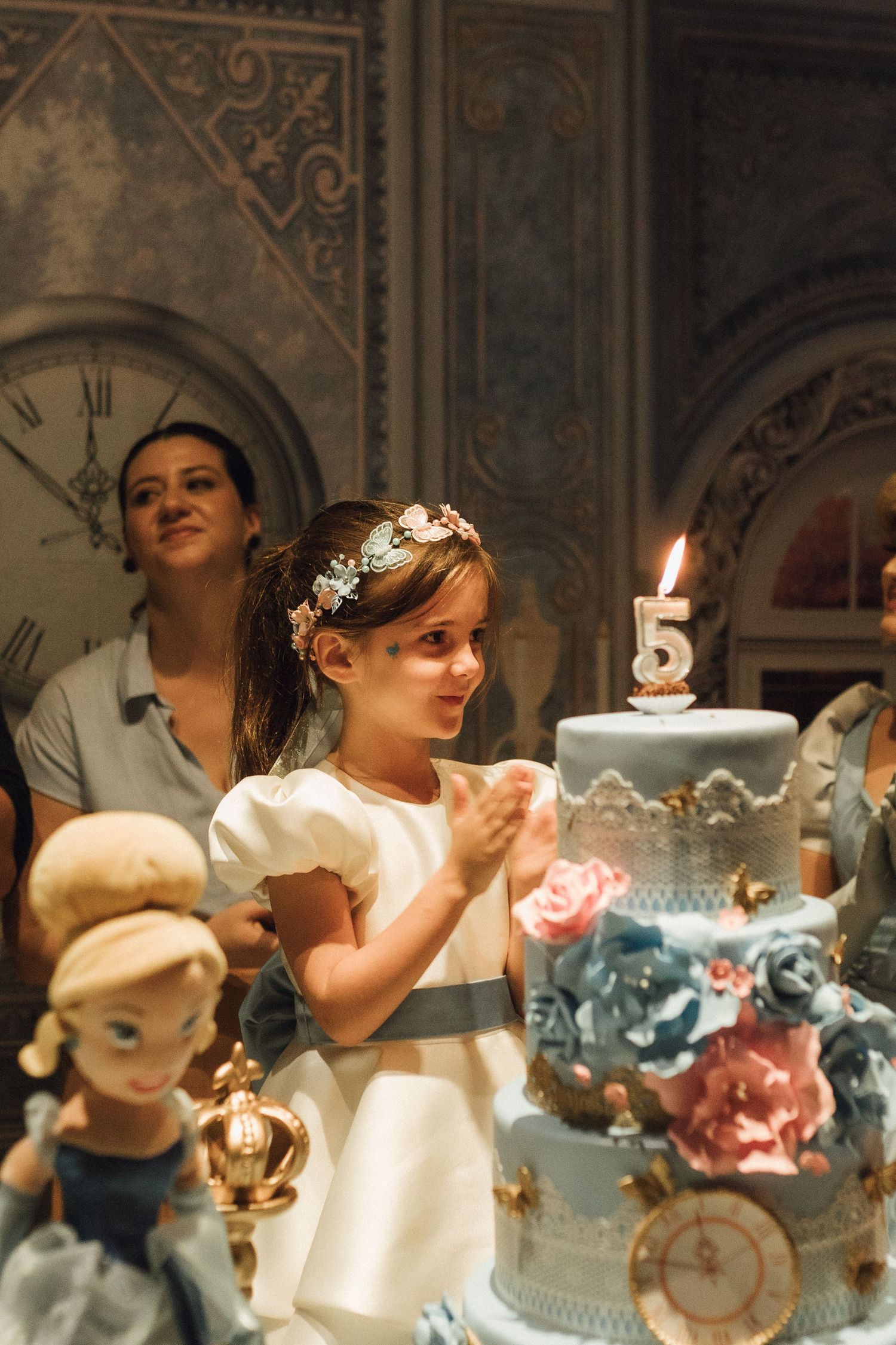 A young girl in a white dress celebrates her 5th birthday with an elegant floral cake and Cinderella doll.