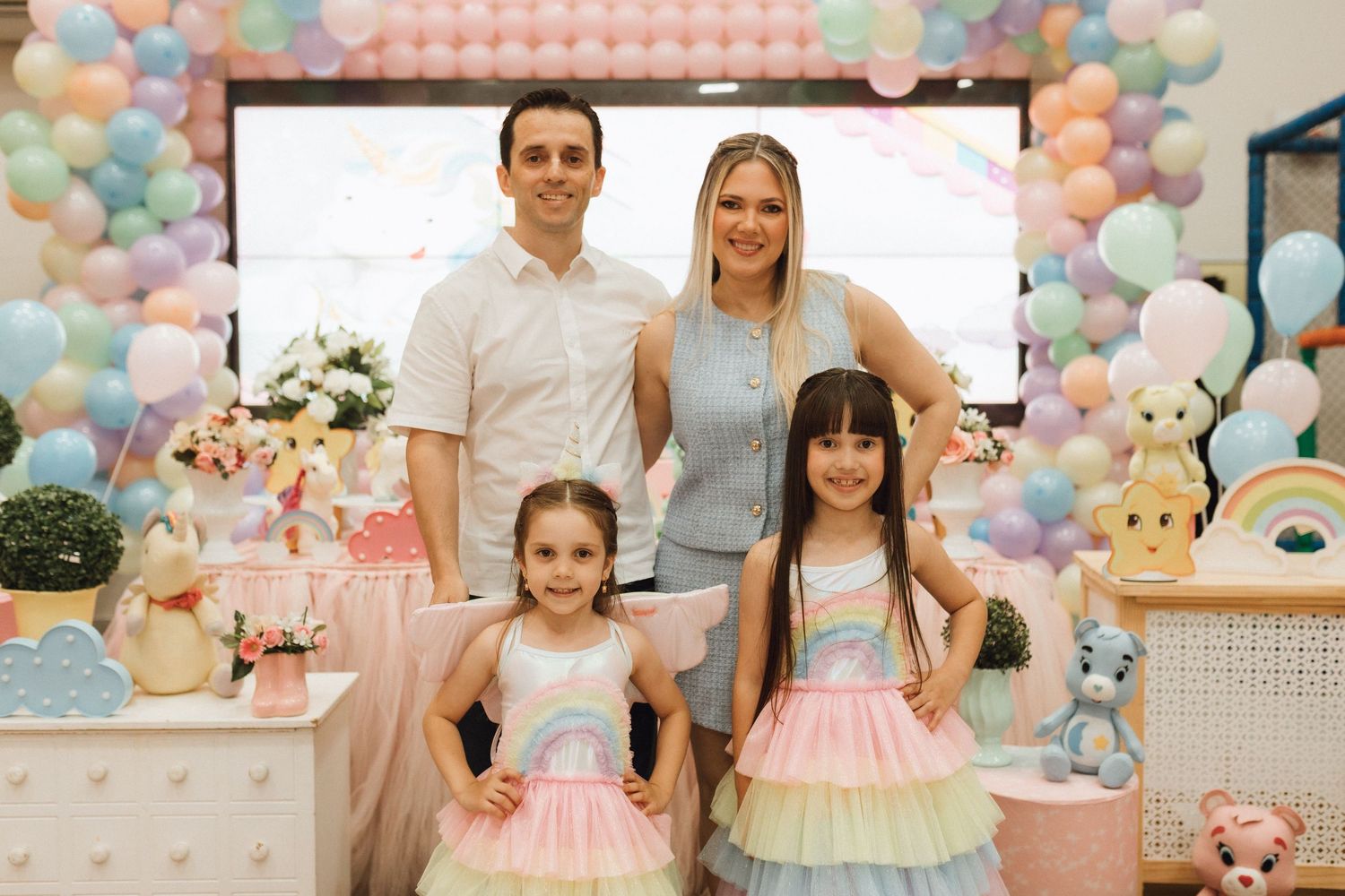 Family of four posing at a pastel rainbow birthday party with balloon arch and decorative table.