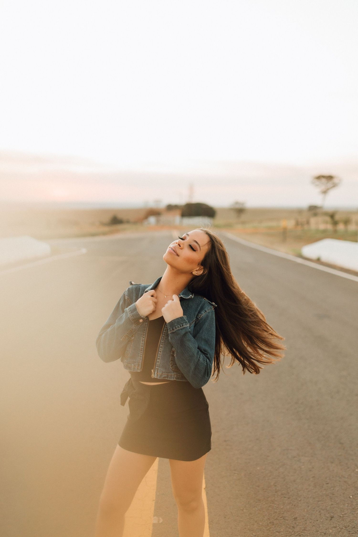 Woman in denim jacket and black skirt posing on open road at golden sunset with hair flowing in wind.