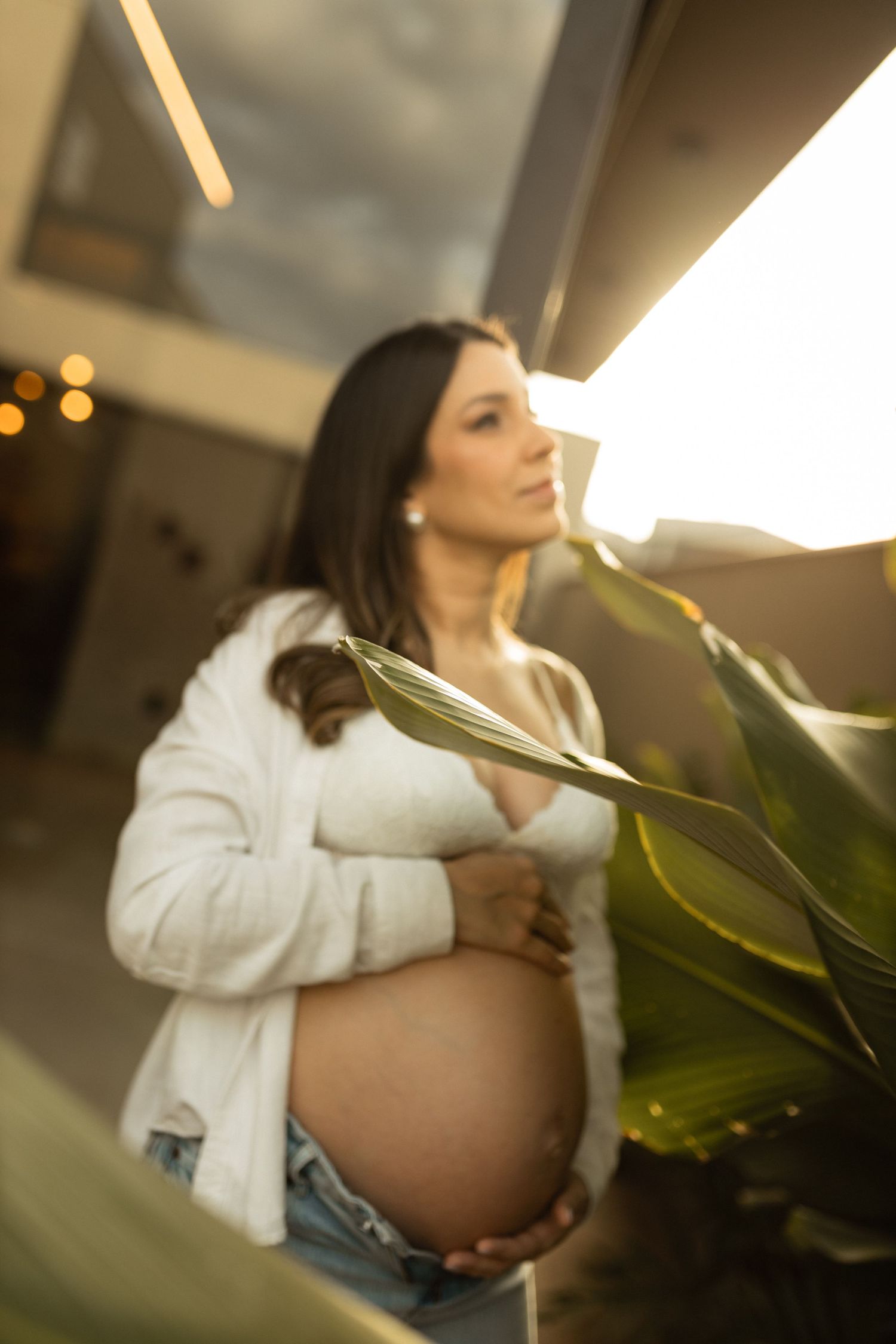 Beautiful pregnant woman in white shirt cradling baby bump beside tropical plant in warm sunset light.