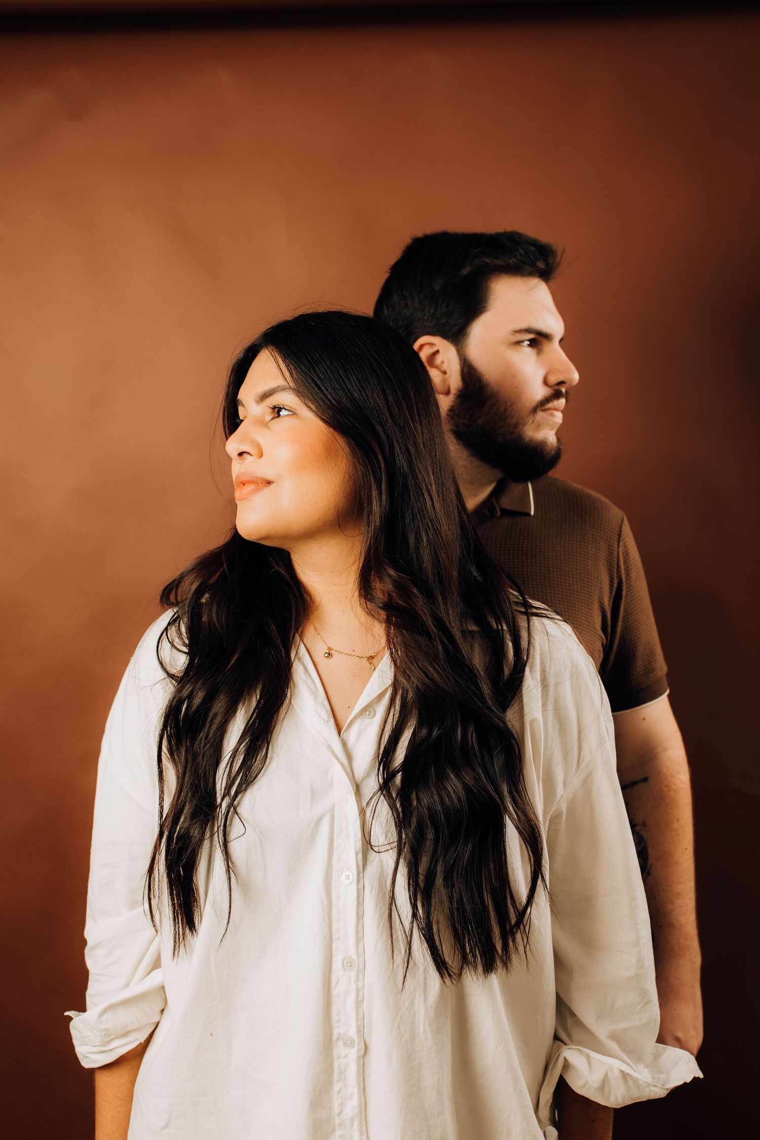A woman and man stand back-to-back in a studio, gazing away, against a warm brown backdrop.
