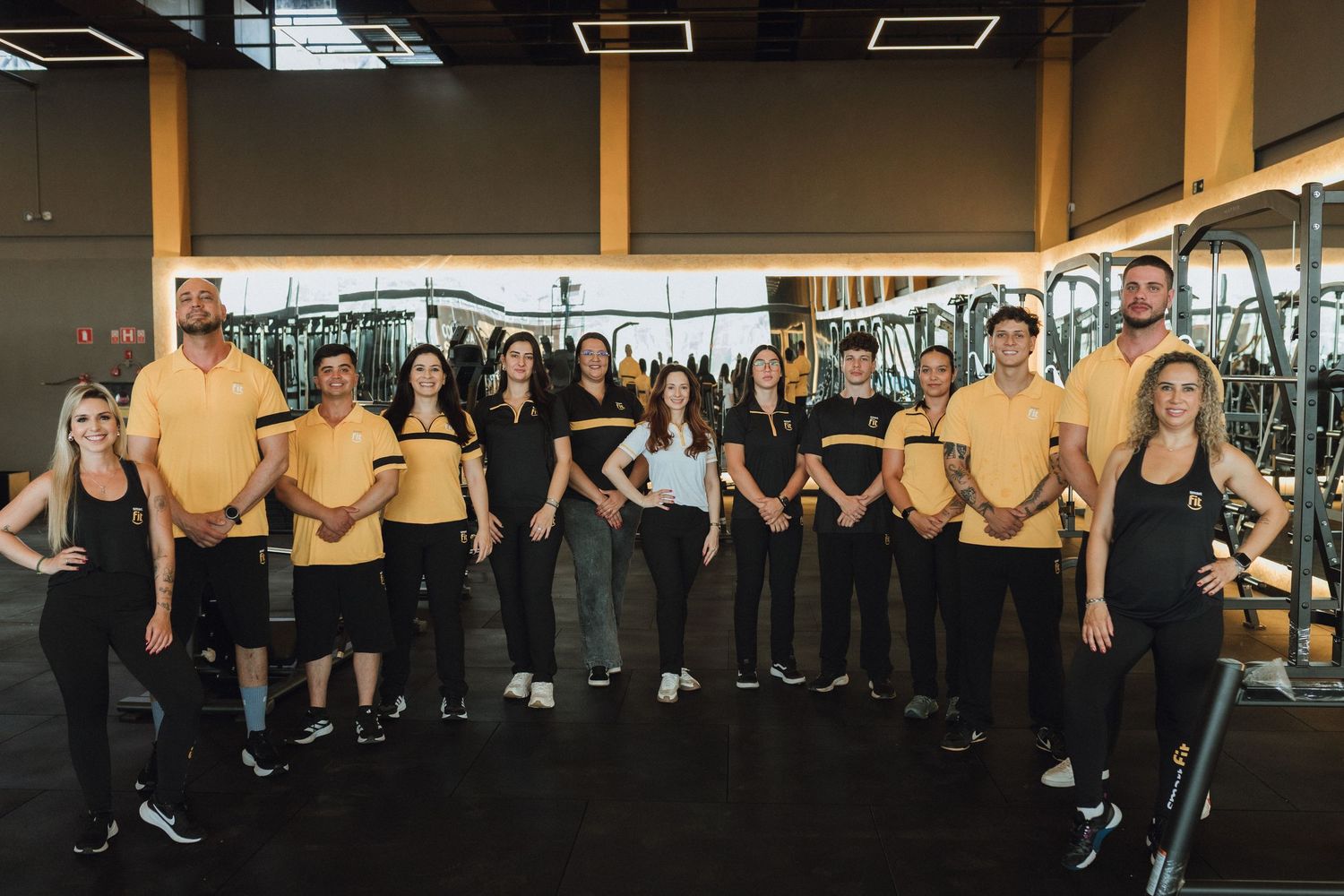 Gym staff team posing together in black and gold uniforms inside a modern fitness center with equipment.