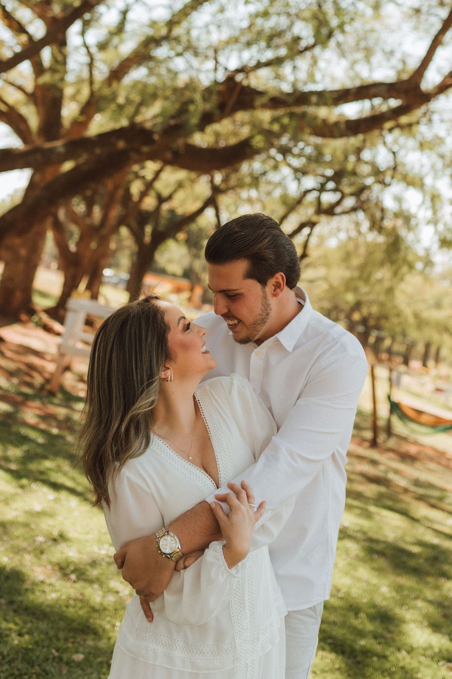 A couple in white outfits embracing and smiling under large trees in a sunlit park setting.