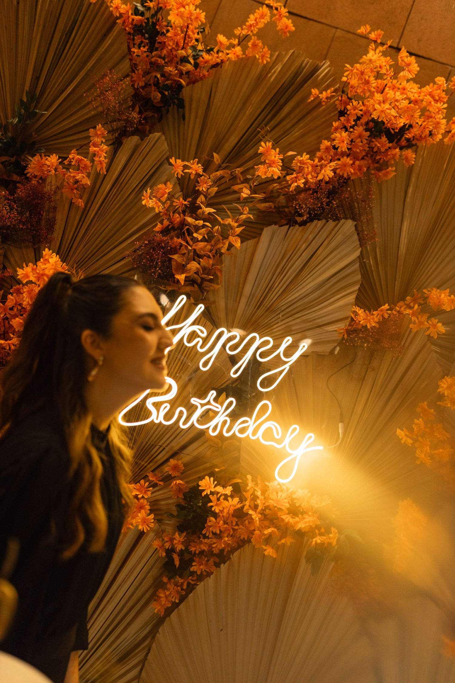 Woman standing beside a glowing Happy Birthday neon sign surrounded by orange flowers and dried palm leaves.