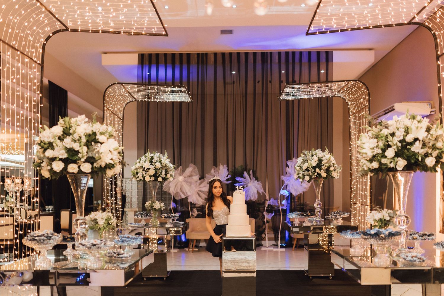 Elegant quinceañera dessert table with white floral arrangements, crystal accents, and fairy lights backdrop.