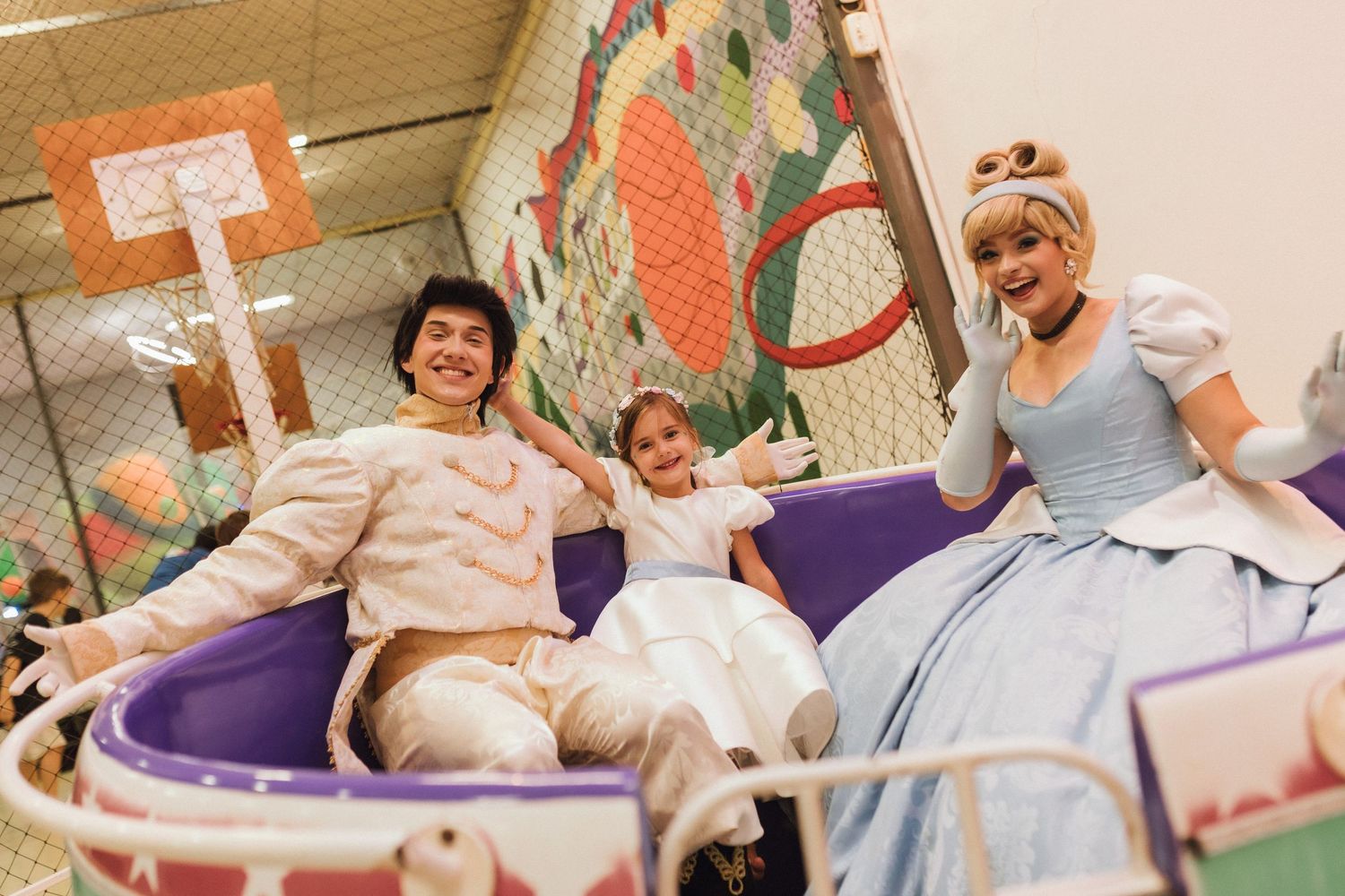 Child riding a teacup ride with Cinderella and Prince Charming characters at a Disney-themed party.