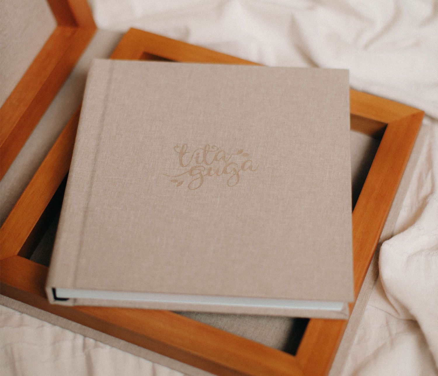 Elegant linen-covered photo album with gold foil script lettering, resting on a wooden frame against white fabric.