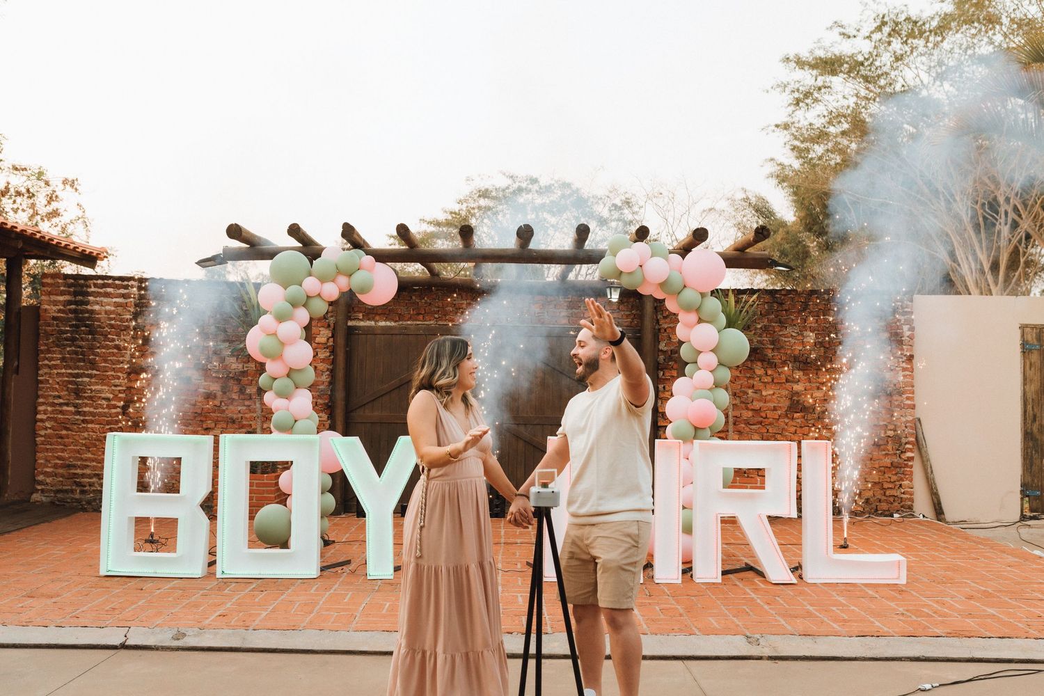 Couple at gender reveal party with pink and blue smoke cannons, BOY GIRL signs, and balloon arch backdrop.