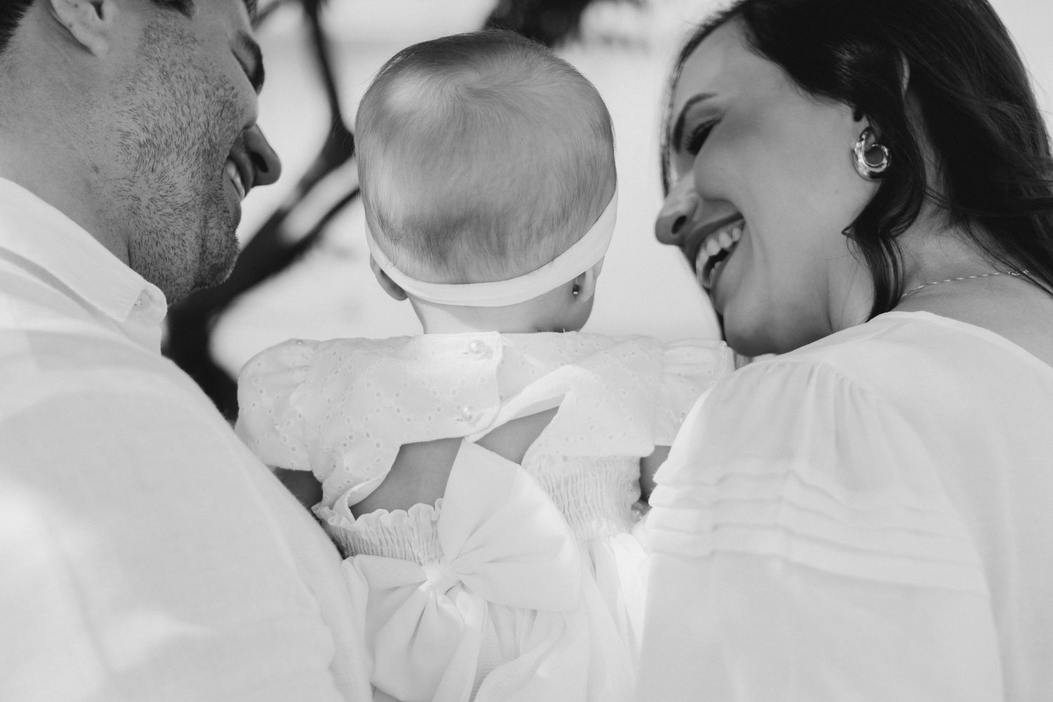 Black and white photo of smiling parents holding their baby dressed in white christening gown.