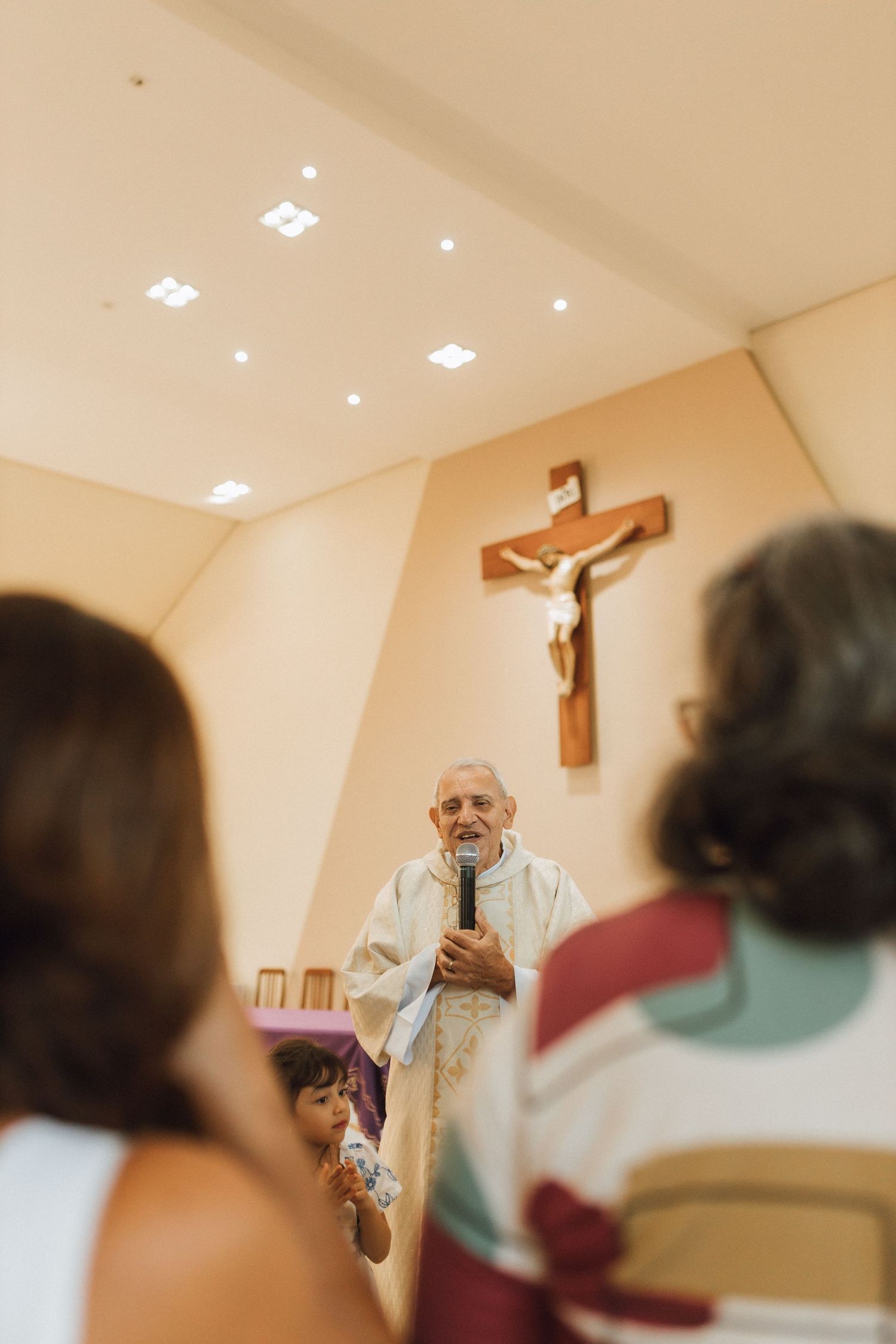 Catholic priest holding microphone speaks at altar beneath crucifix during baptism ceremony.