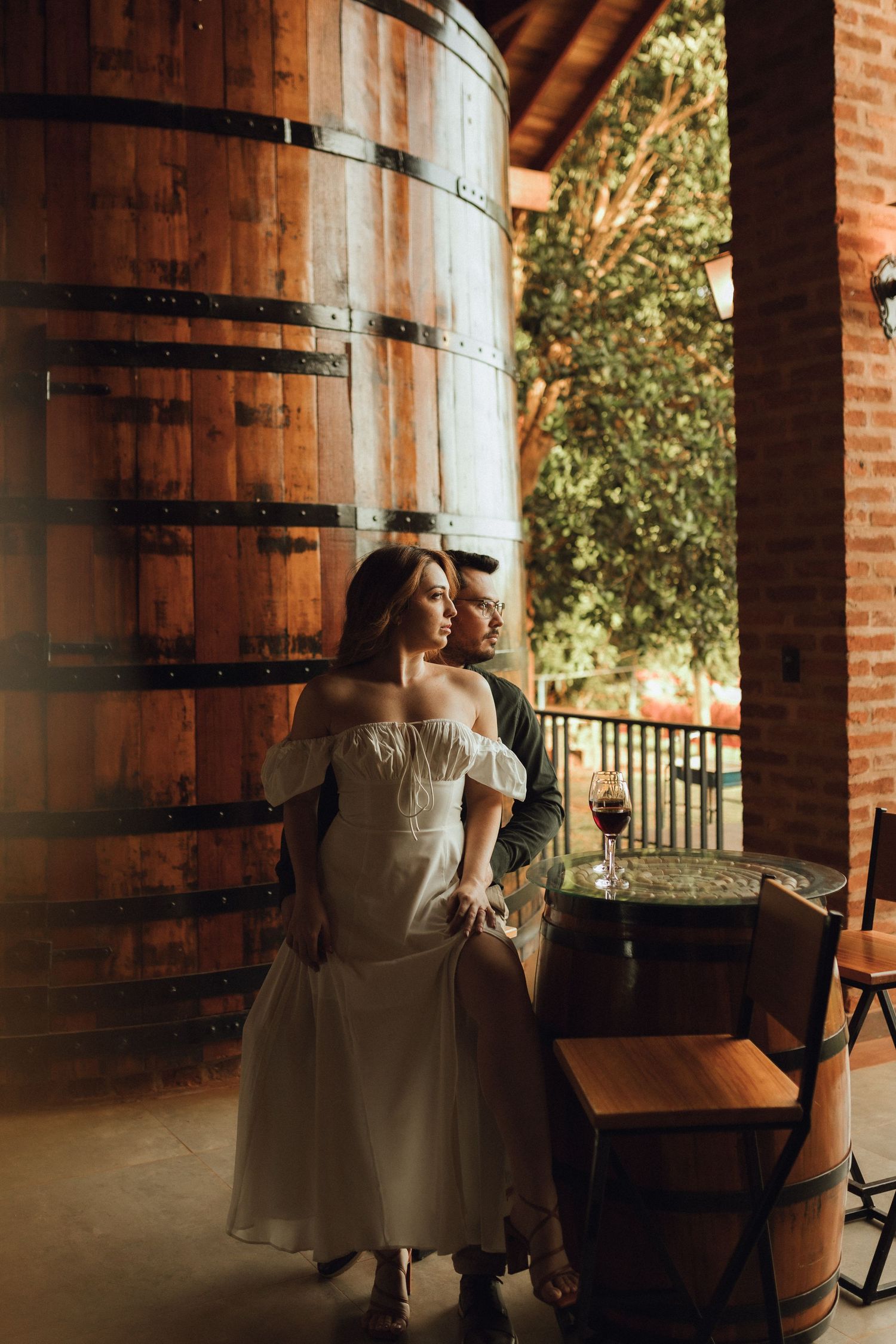 Couple embracing near large wooden wine barrels in a rustic winery, woman holding a glass of red wine.