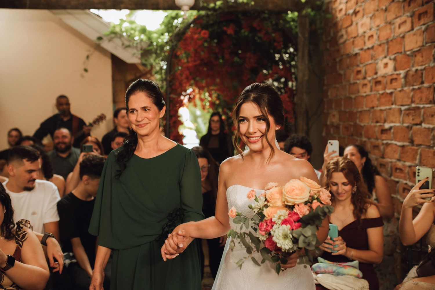 Bride walks down the aisle holding colorful bouquet with her mother at an outdoor rustic wedding ceremony.