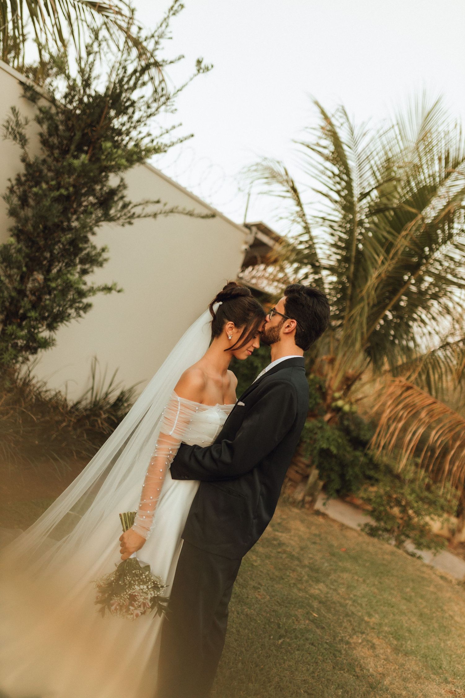 Newlyweds sharing a romantic embrace in garden with palm trees at golden hour.