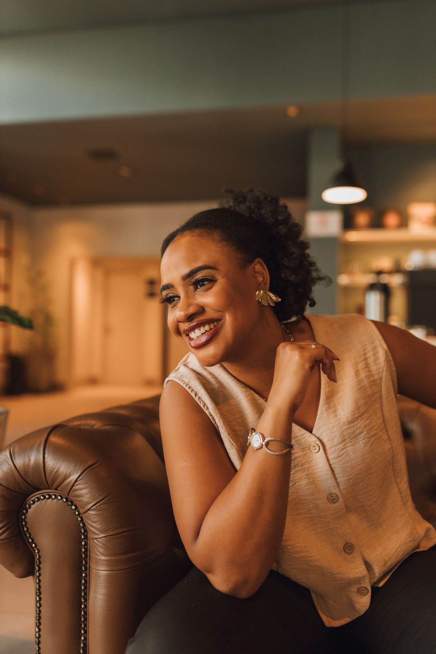 Happy Black woman in beige top smiling while seated in leather armchair indoors.