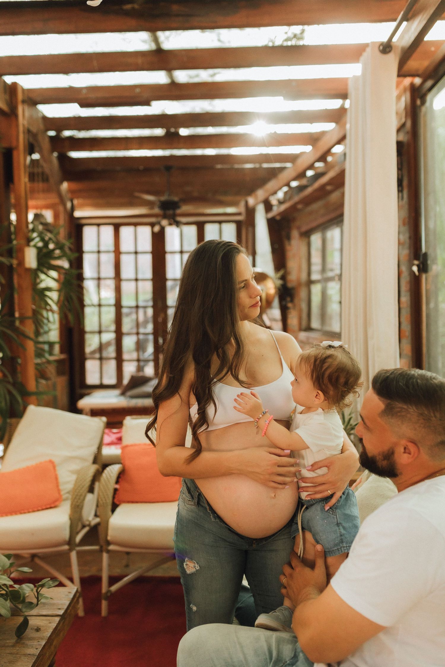 Pregnant woman holding toddler with husband beside her in a warmly lit sunroom setting.