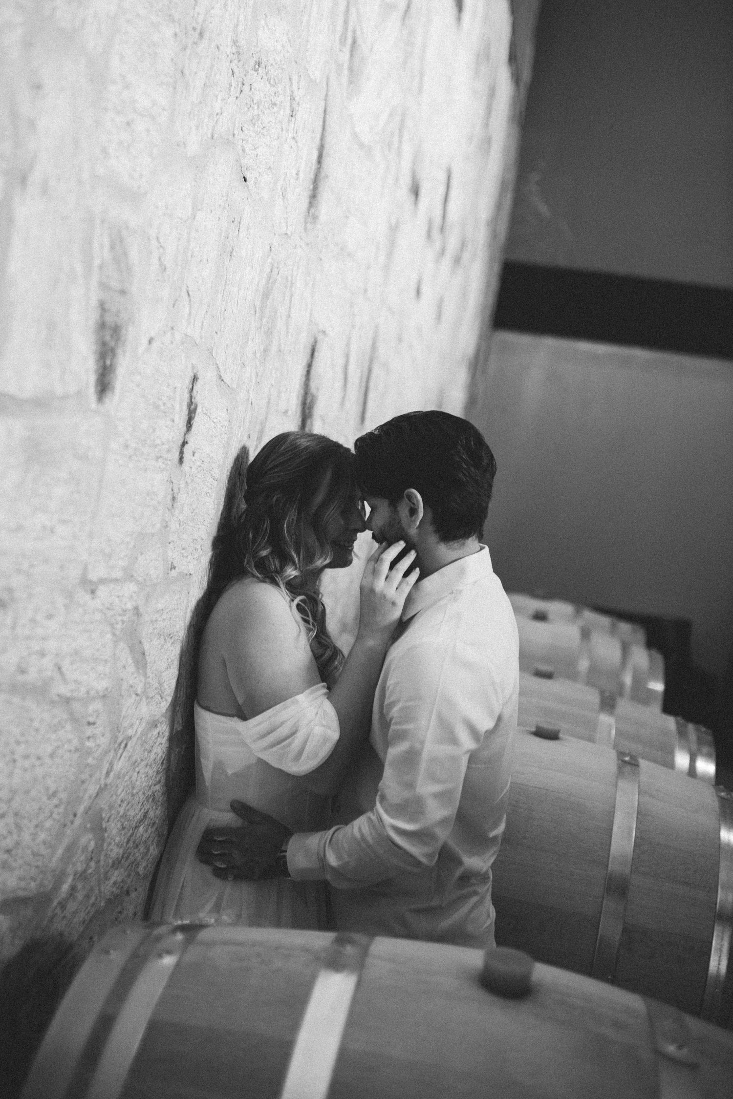 Black and white photo of a romantic couple sharing an intimate moment leaning against a stone wall surrounded by wine barrels.
