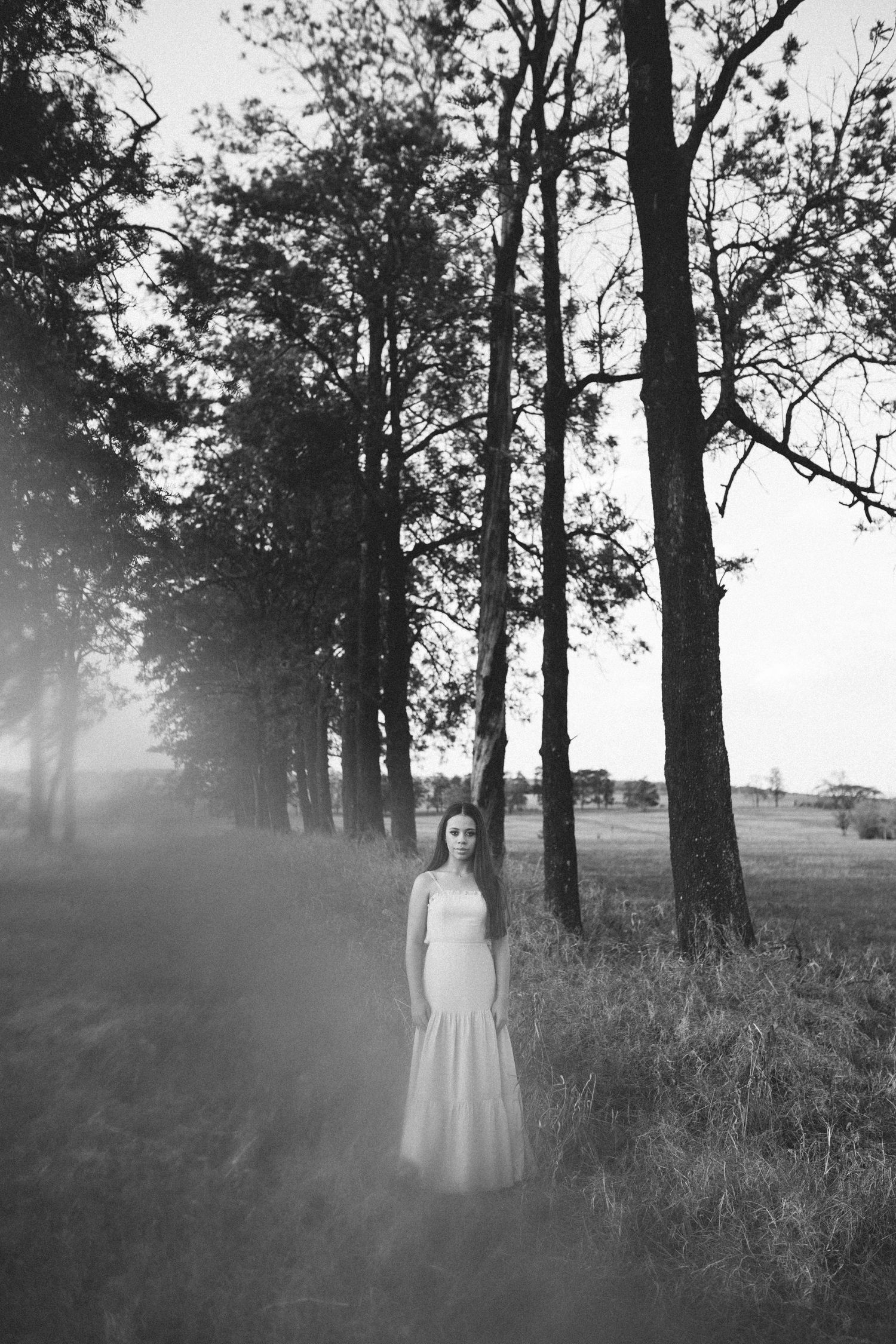 Black and white photo of a woman in a white dress standing among tall trees in a misty field at dusk.