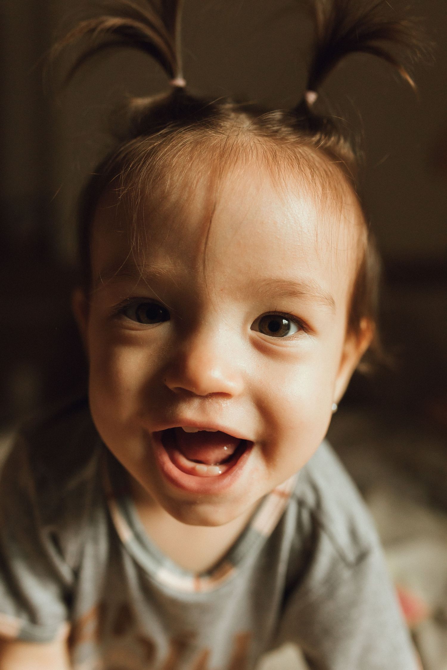 Smiling toddler girl with two small pigtails wearing a gray shirt, showing her baby teeth in a joyful expression.