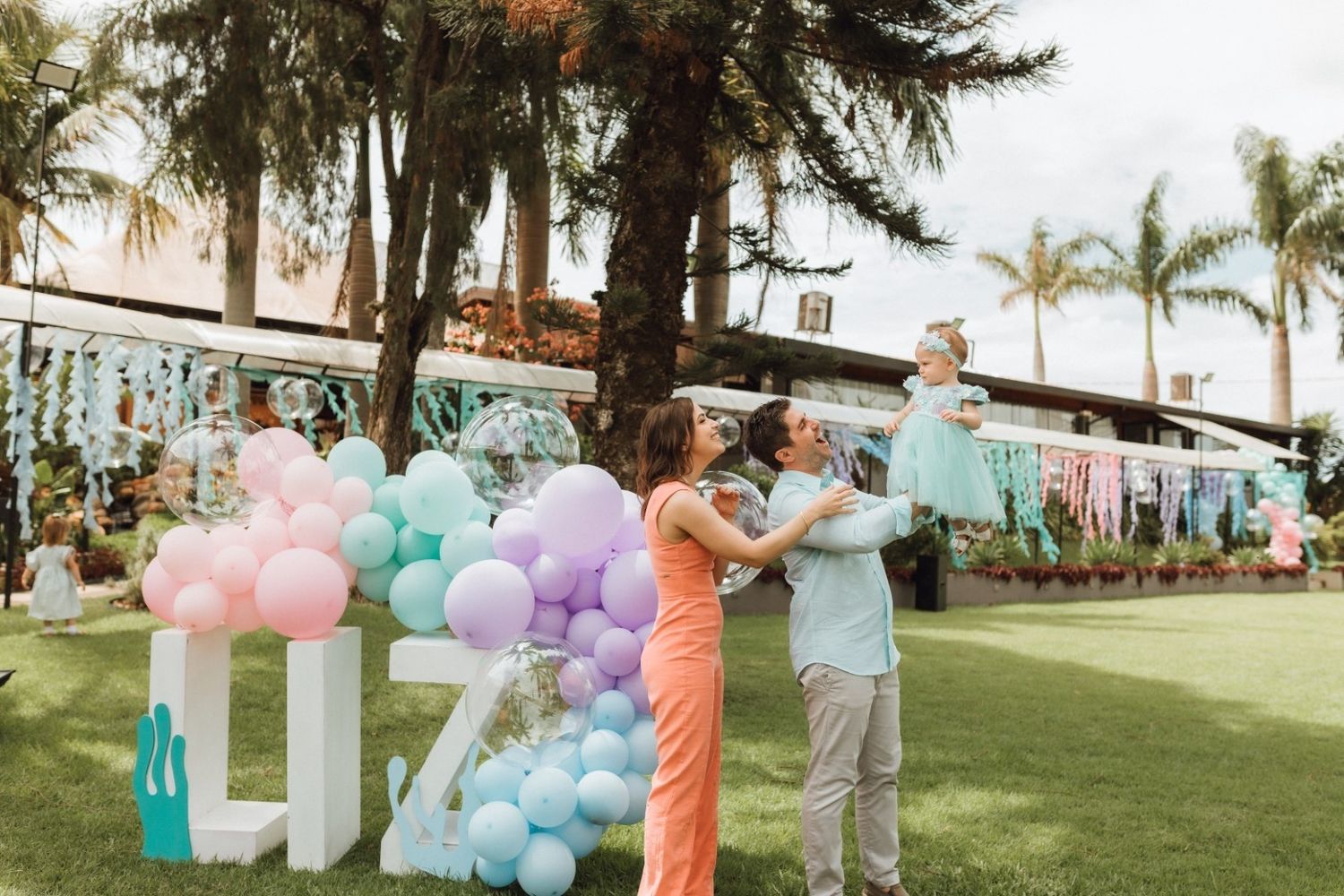 Family celebrating a gender reveal party with colorful balloons and pastel decorations on a tropical lawn.