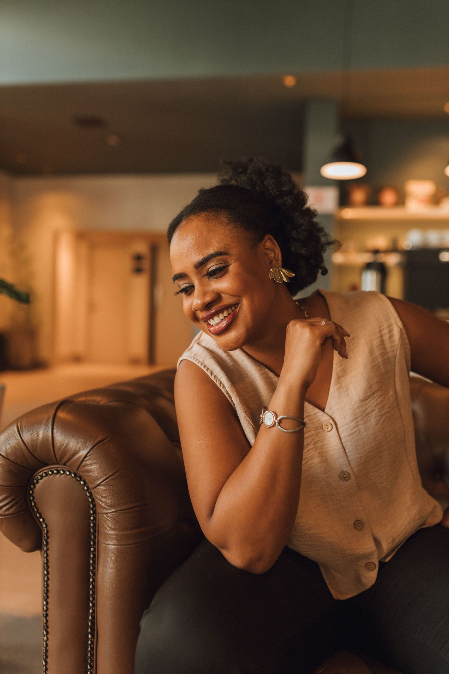 Smiling young Black woman relaxing on brown leather sofa in warmly lit modern interior space.
