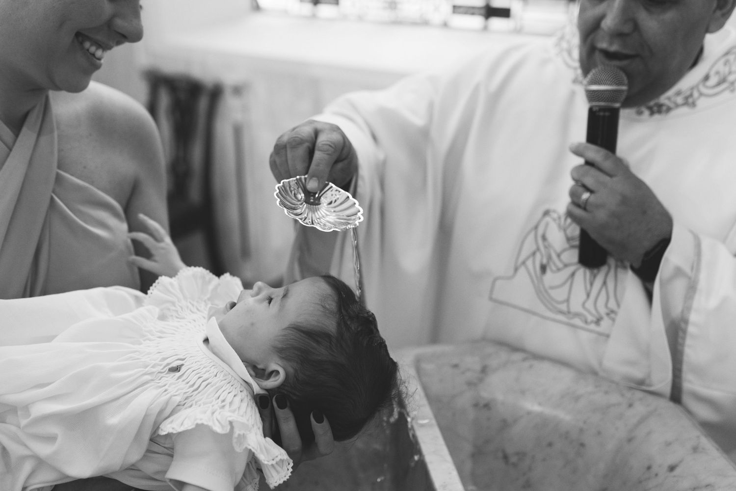 Priest performing a Catholic baptism ceremony, pouring holy water over a newborn baby in white christening gown.