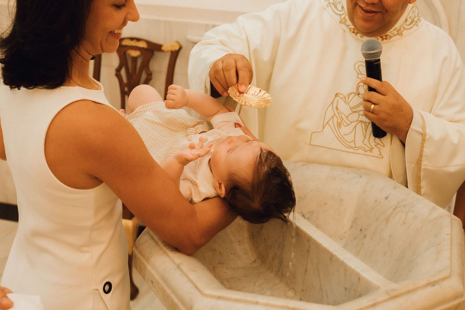 Priest performing a Catholic baptism, pouring holy water over a newborn baby held by the mother.