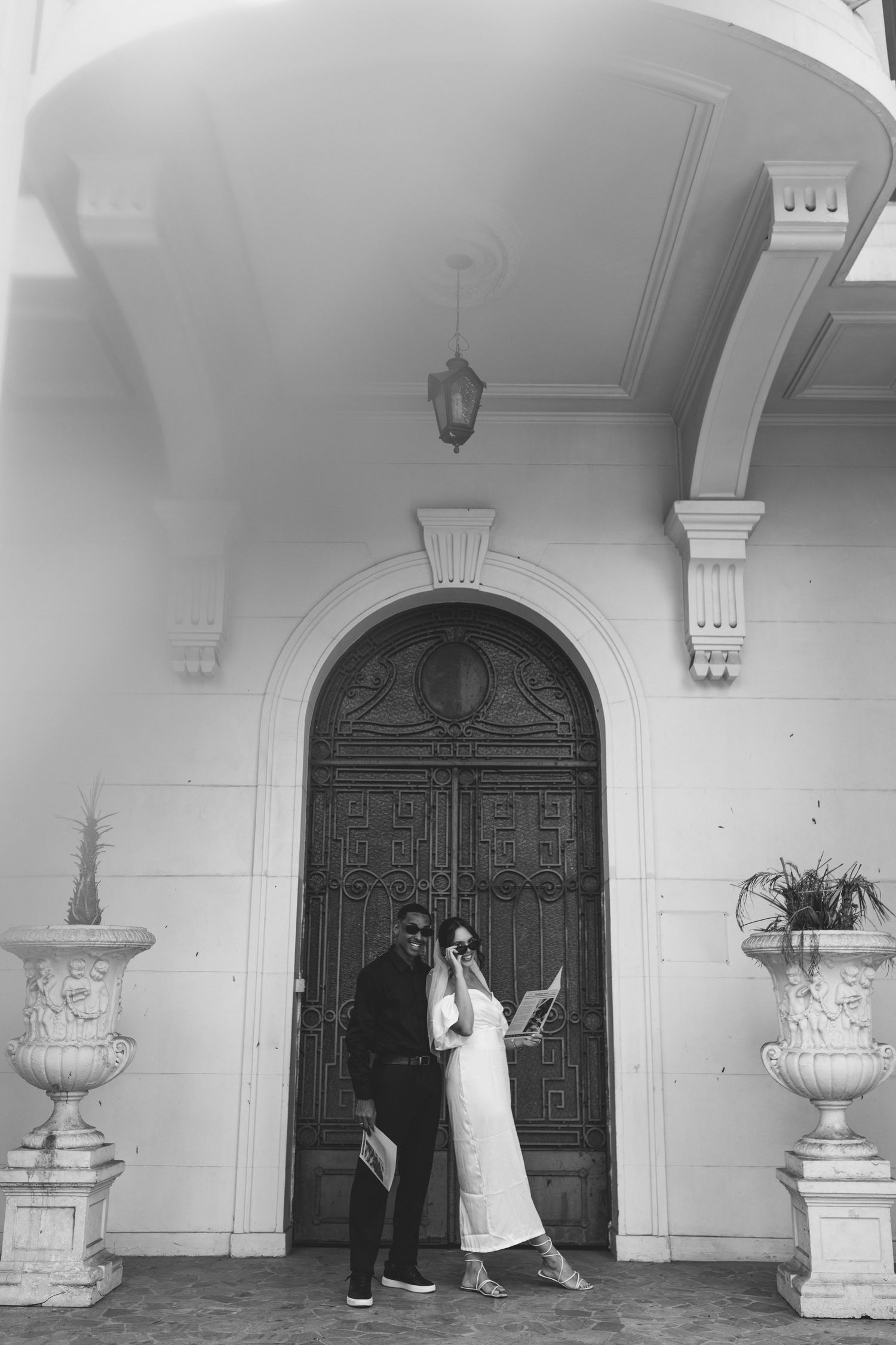 Couple posing together in front of elegant arched wooden door flanked by classical stone urns, black and white.
