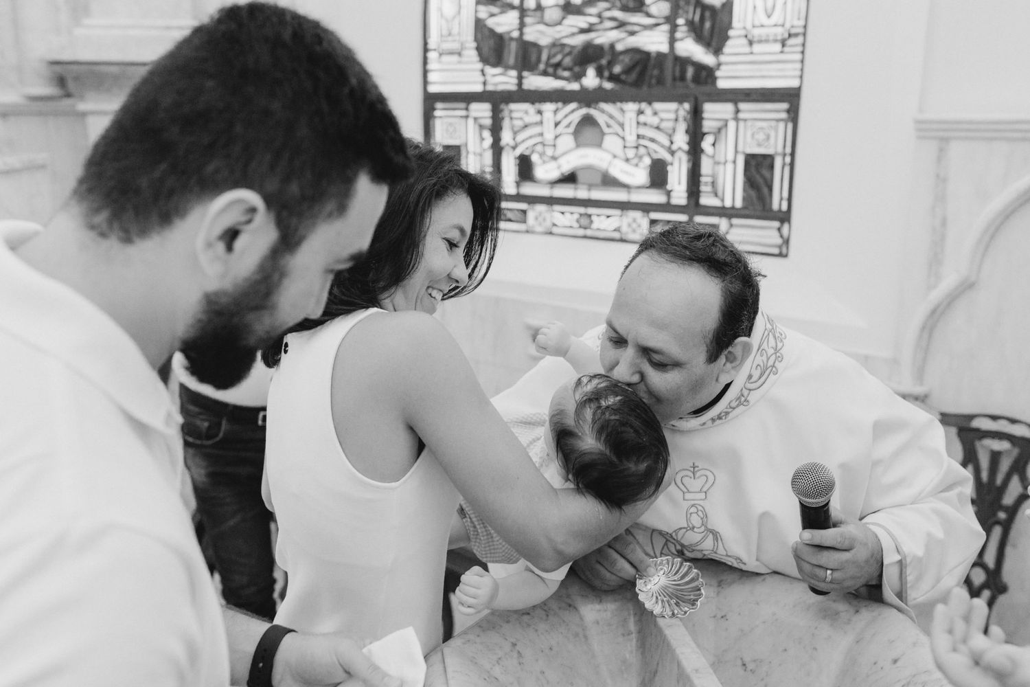 Black and white photo of a priest baptizing a baby while parents watch in a church ceremony.