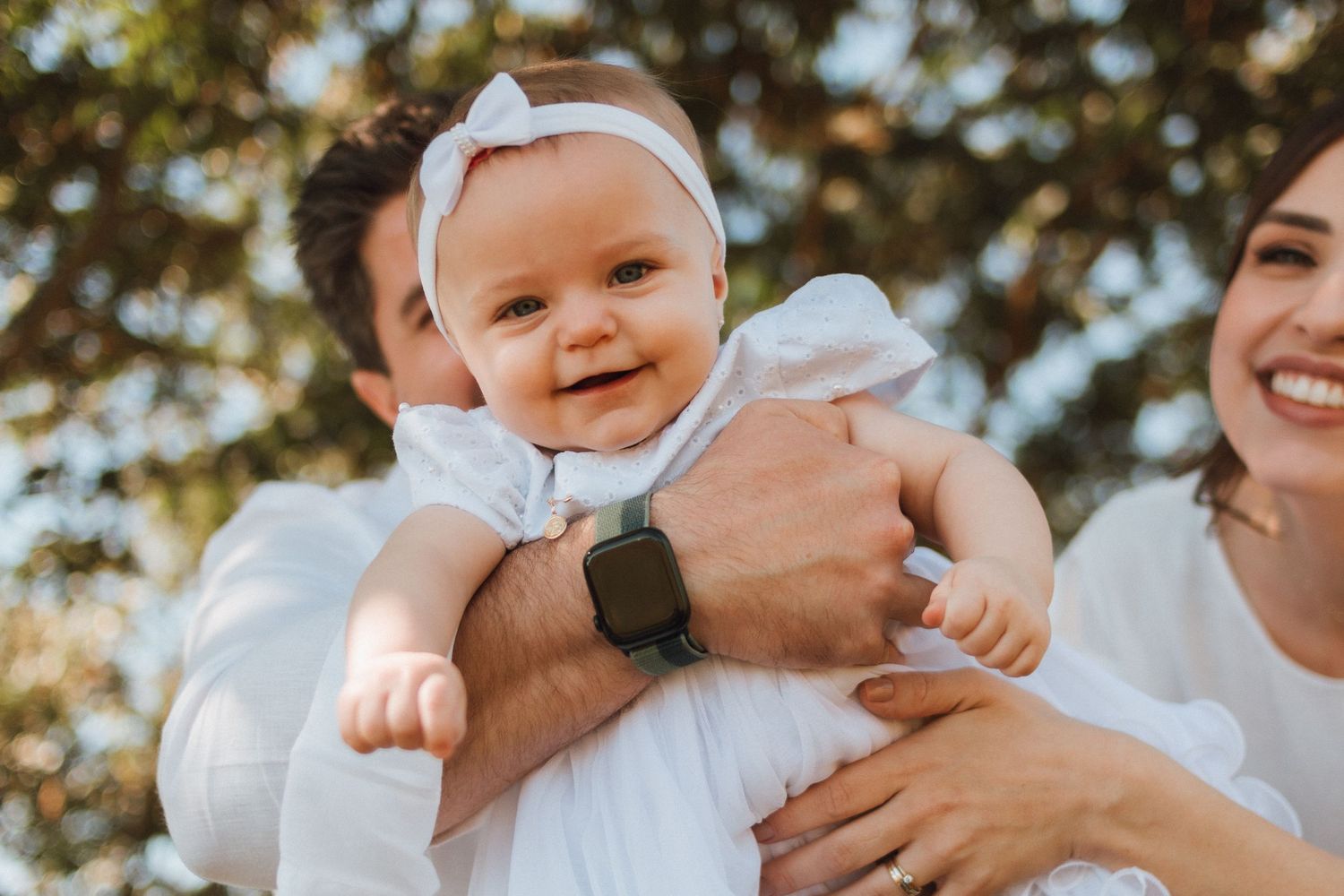 Happy baby girl in white dress with bow headband held by parents outdoors in a sunny park setting.