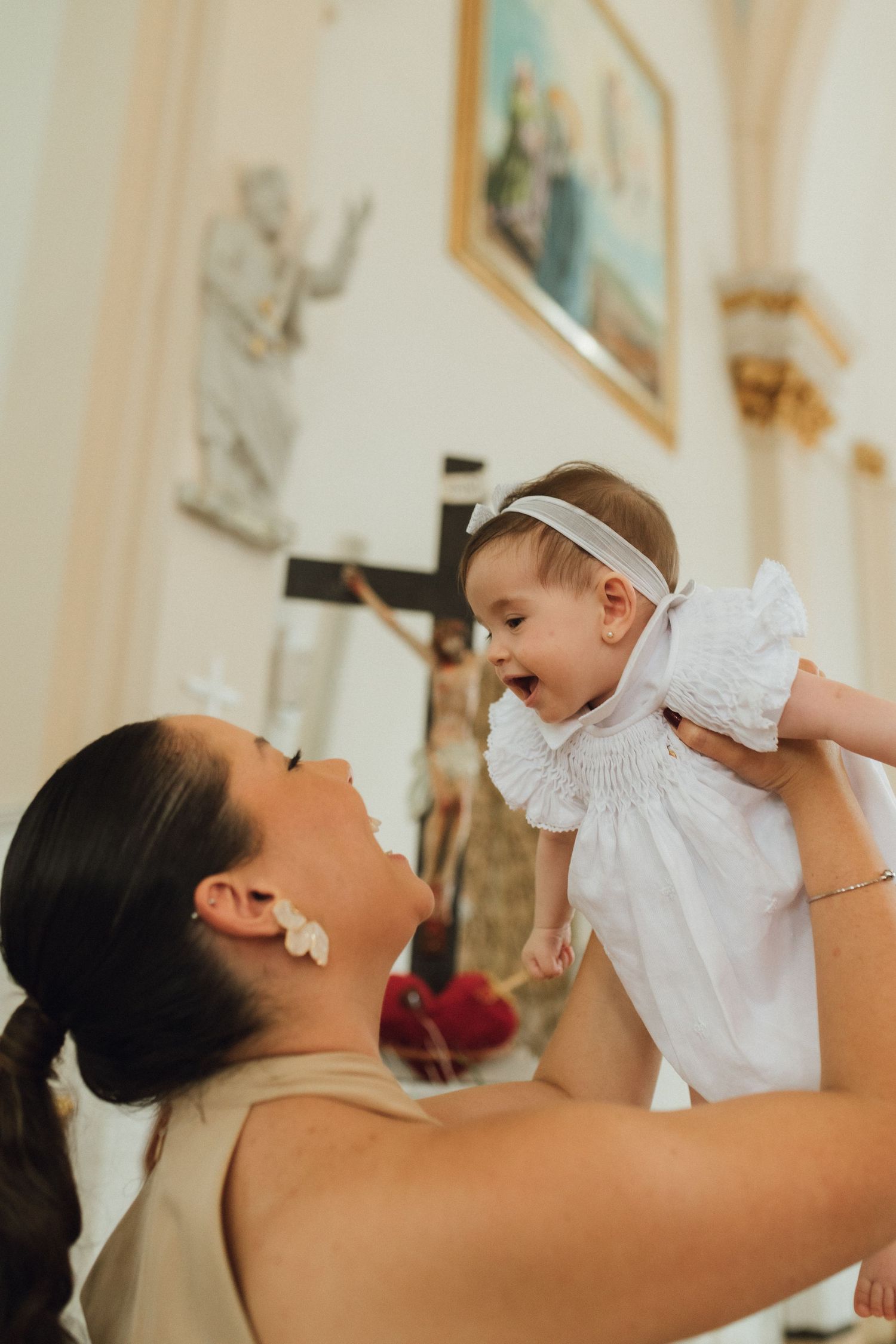 Joyful mother holding smiling baby girl in white dress at Catholic church baptism ceremony.