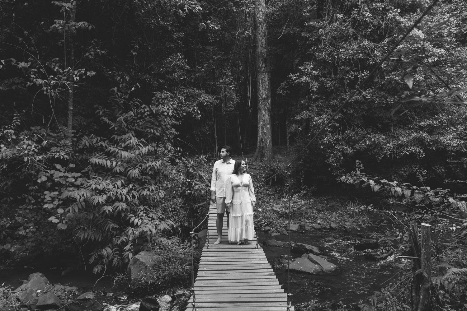 Black and white photo of a couple standing on a wooden bridge surrounded by lush forest trees.
