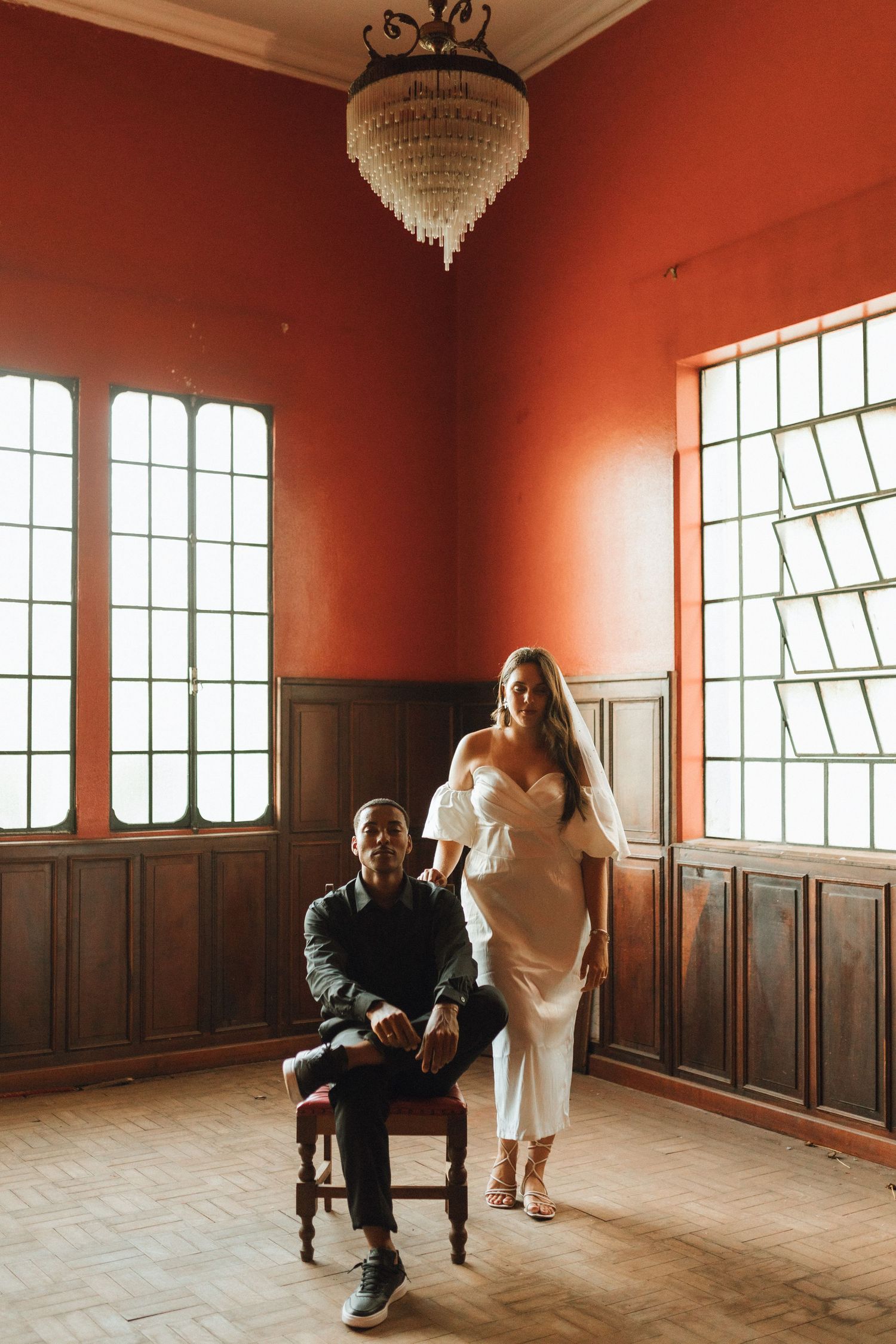 Bride in white dress and groom in dark suit pose in a red-walled room with chandelier and tall windows.