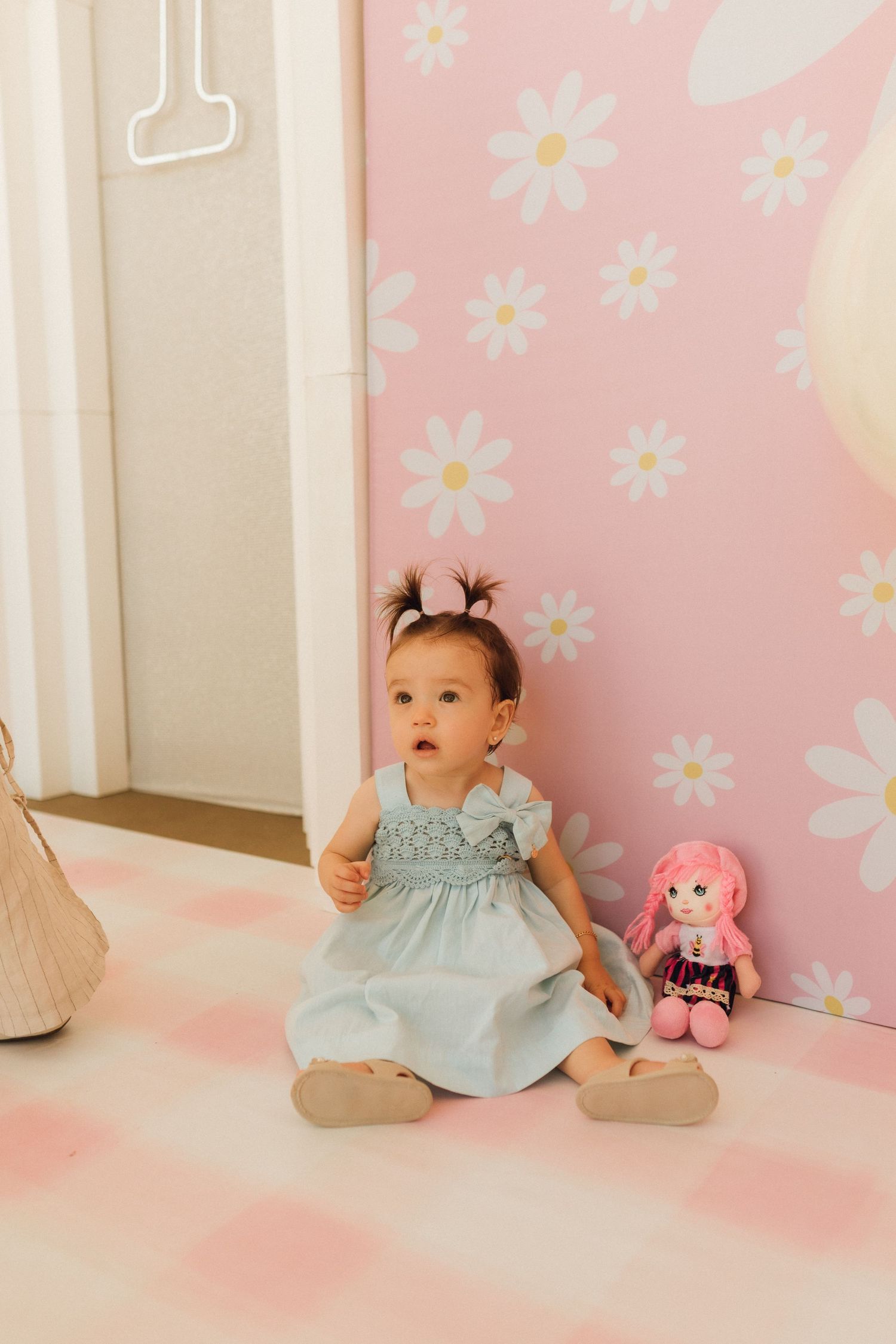 Baby girl in blue dress sits on pink floral mat beside a doll, against a daisy-print pink wall.