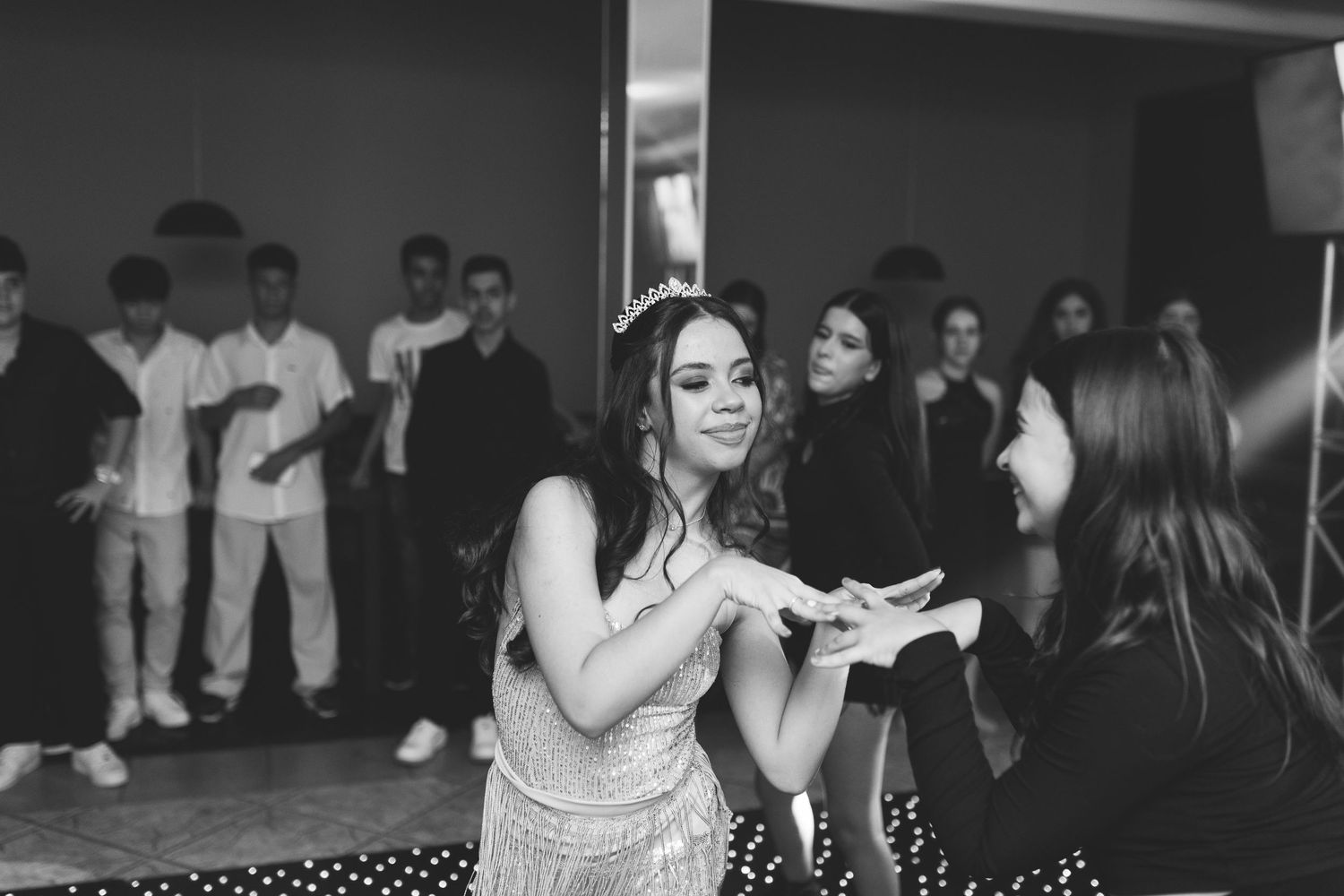 A young woman wearing a tiara dances at a party while guests watch in the background.