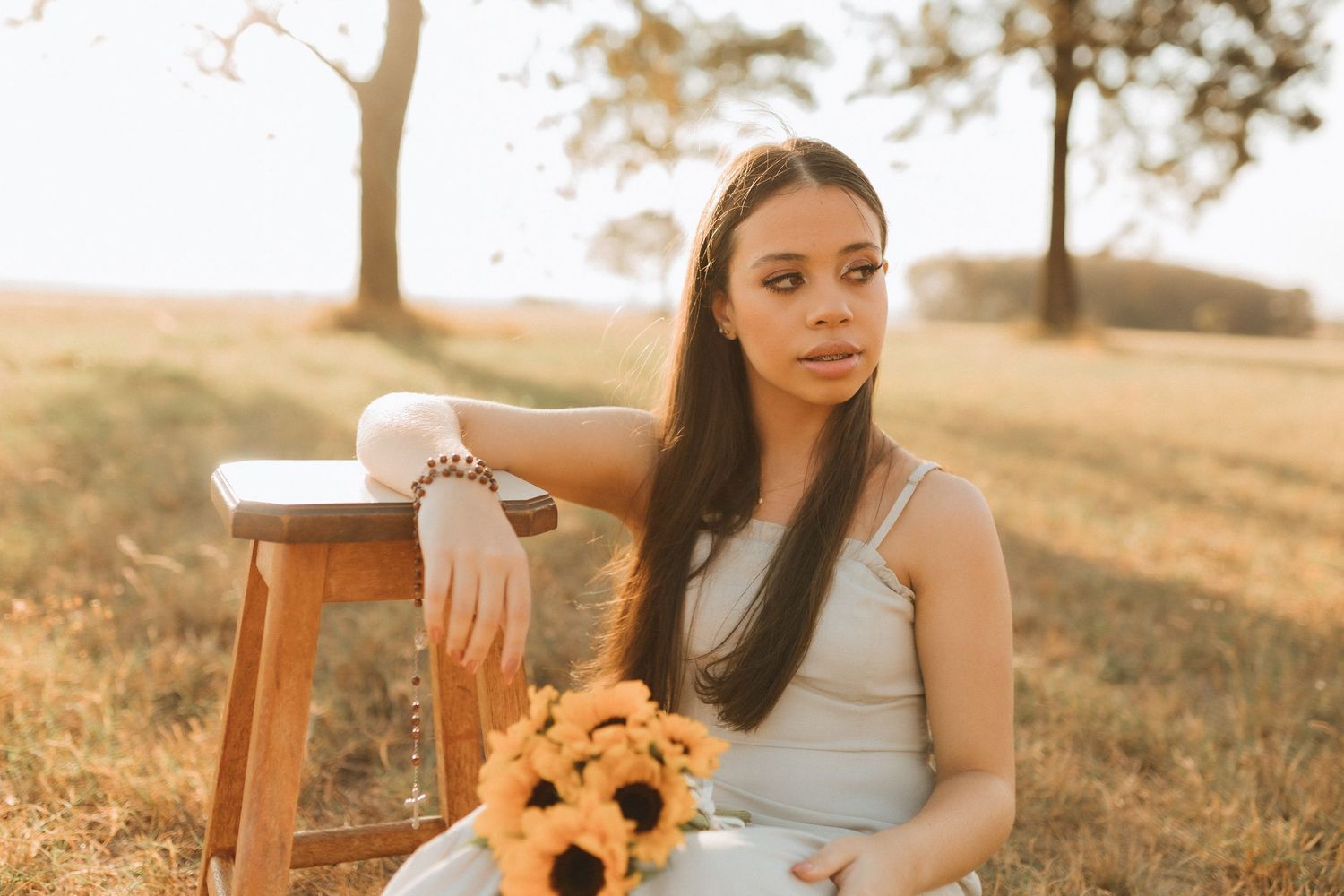 Young woman in white dress holding sunflowers sits beside wooden stool in golden sunlit field.