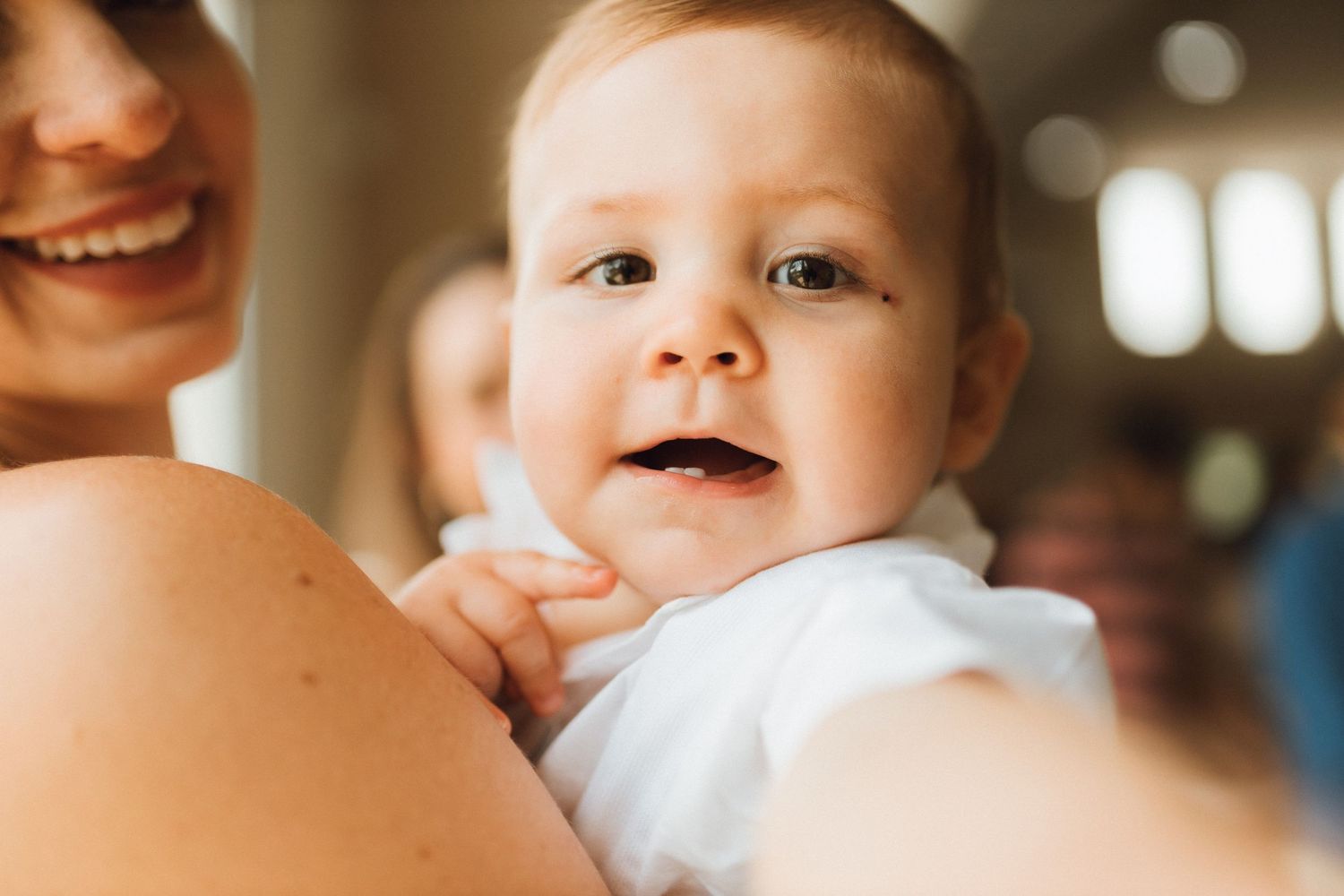 Happy baby in white outfit being held by smiling mother, looking at camera with bright eyes and open mouth.