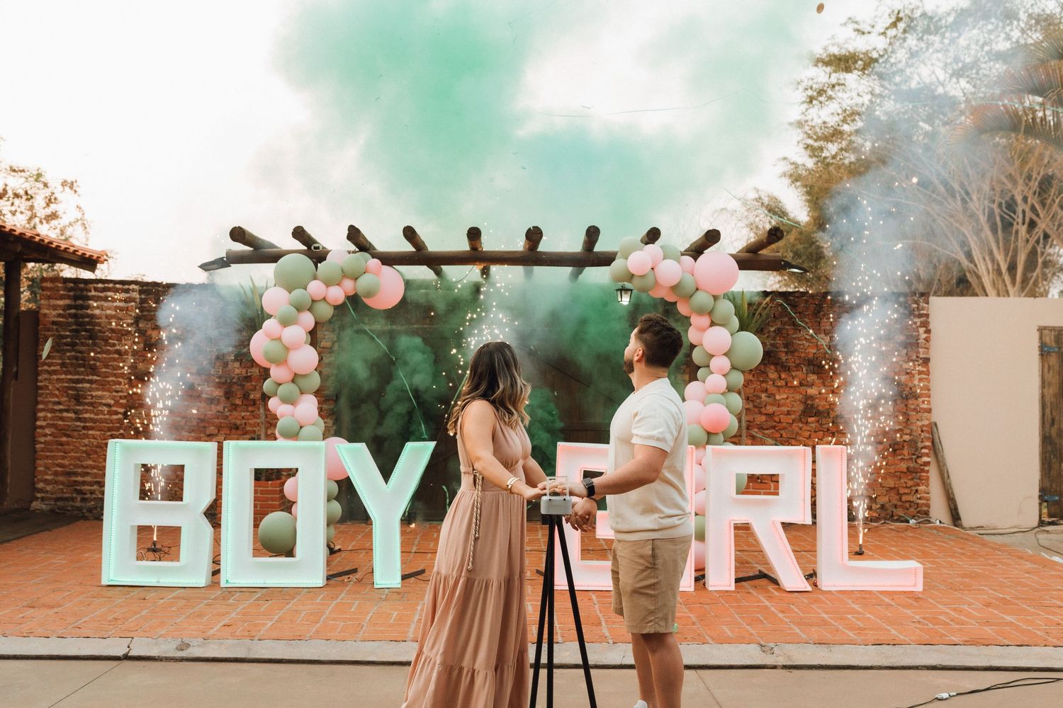 Gender reveal party with BOY GIRL signs, balloon arch, and colorful smoke cannons outdoors.