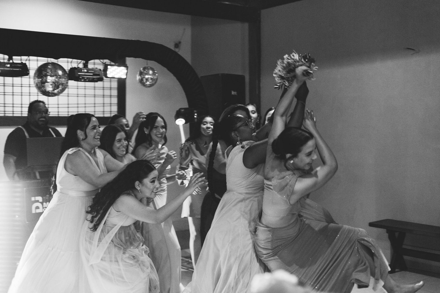 Black and white photo of excited guests competing to catch bridal bouquet at reception.