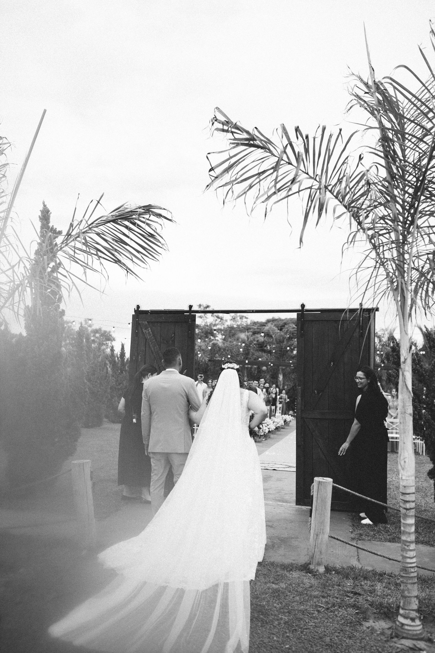 Black and white photo of bride in long veil walking through barn doors with groom at outdoor wedding.