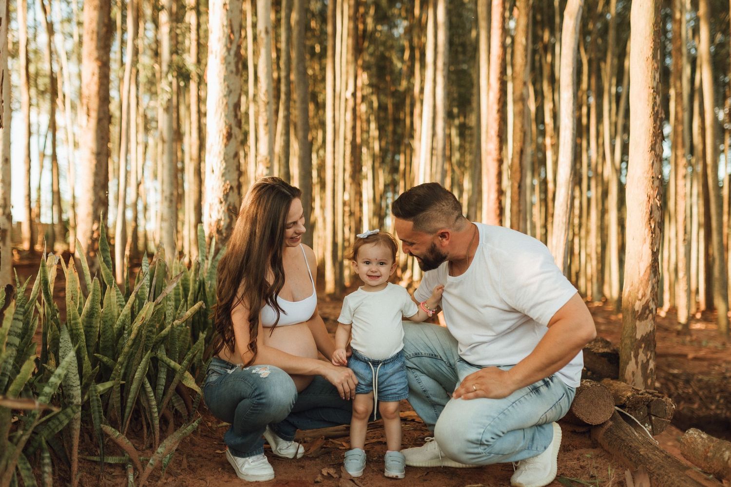 Happy family with toddler posing in a sunlit pine forest, parents smiling at child in a warm outdoor photo.