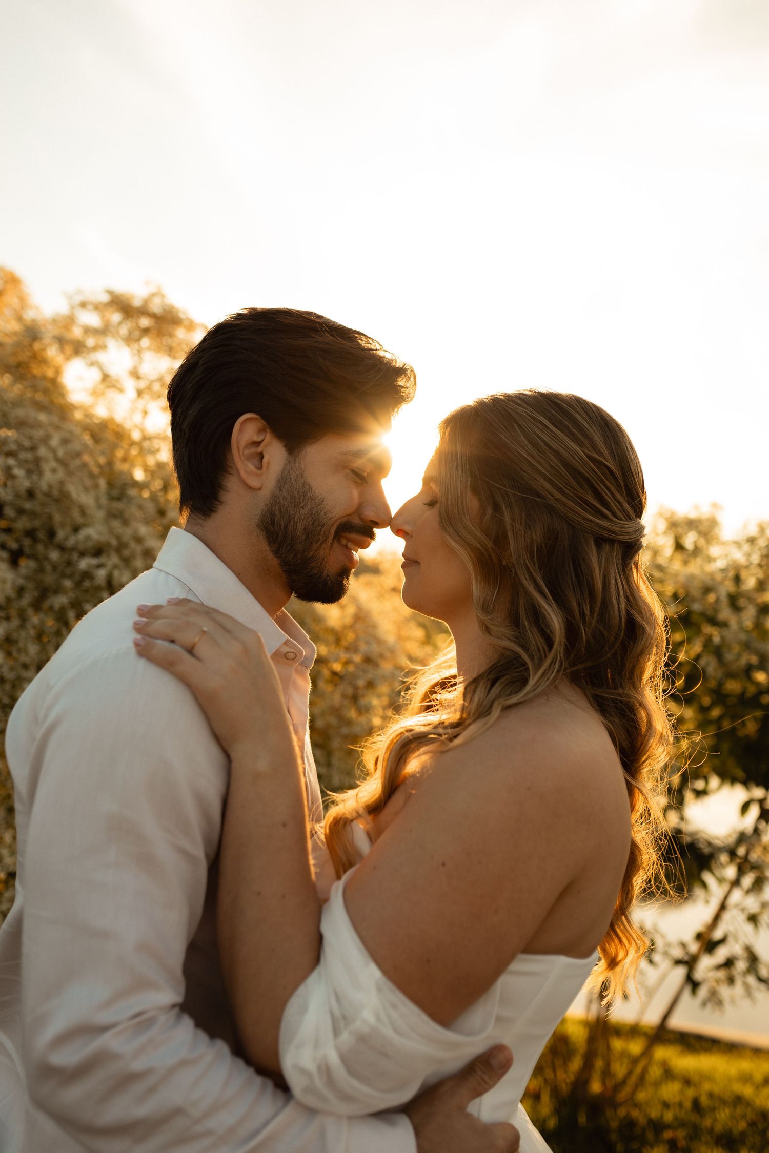 Couple sharing a romantic moment at golden hour sunset, smiling and embracing outdoors near trees.