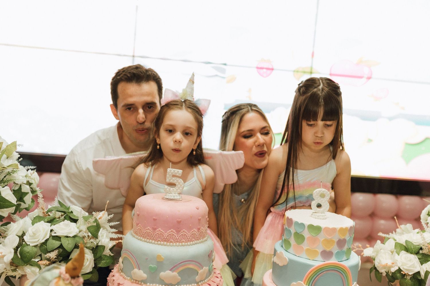 Family of four celebrating a birthday with unicorn cakes, two girls wearing party hats blowing out a number five candle.